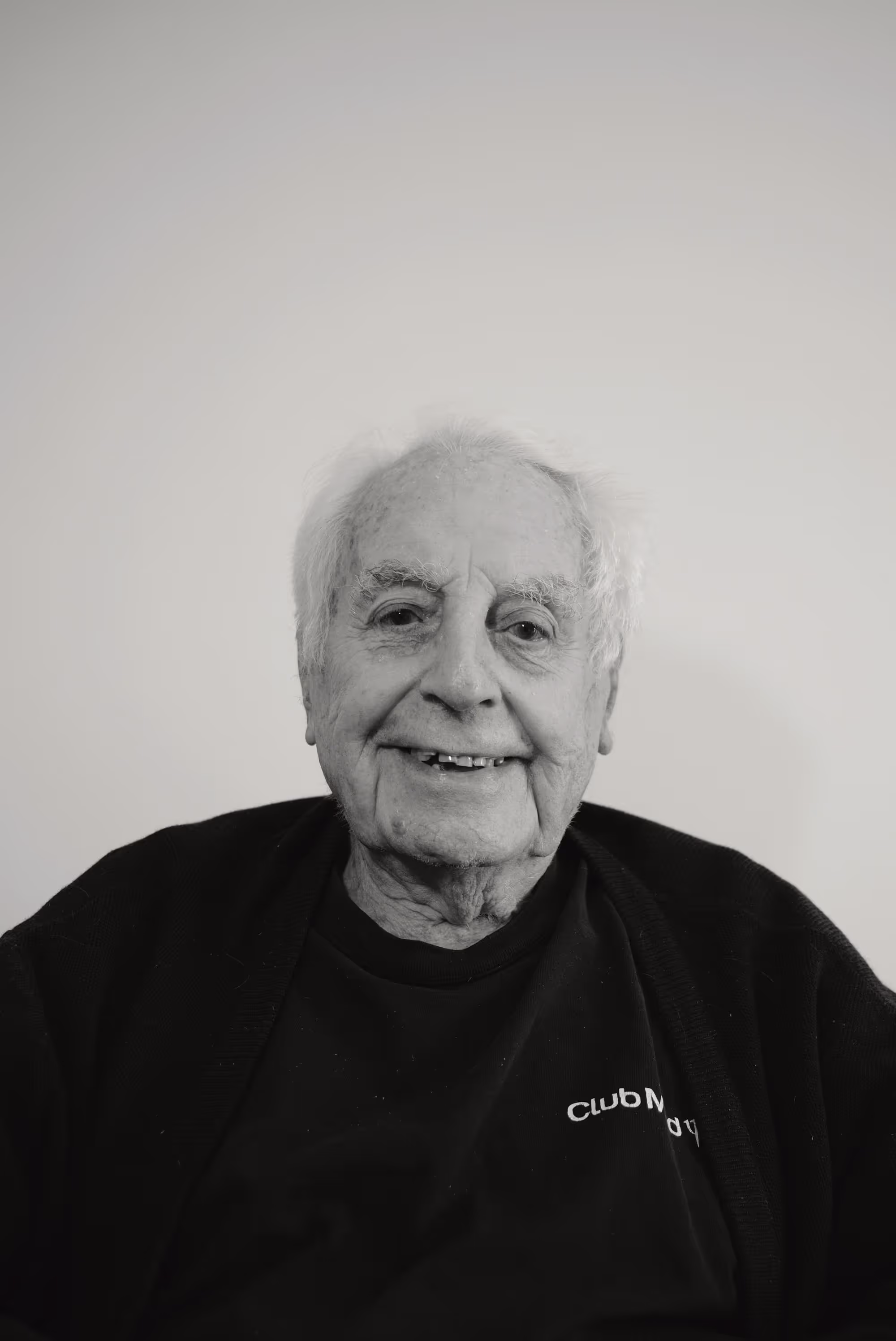 Portrait souriant d’un homme âgé vêtu d’un cardigan et d’un t-shirt noir, sur fond clair.