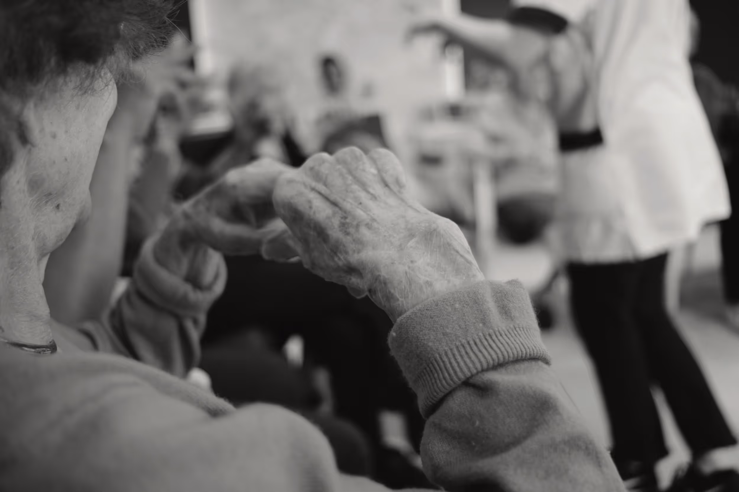 Mains ridées jointes au premier plan, personnes âgées en arrière-plan dans une activité collective.