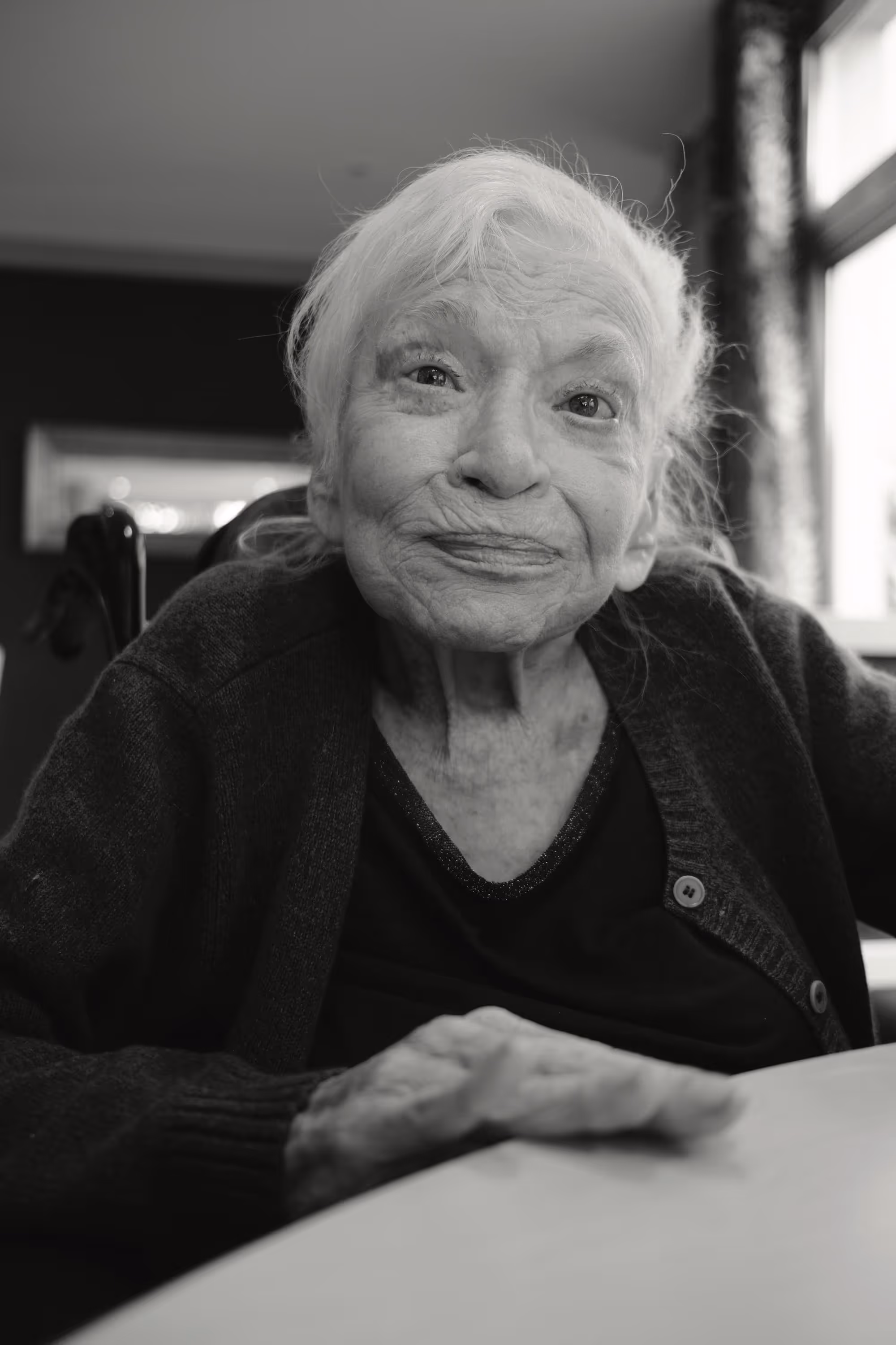 Portrait d'une femme âgée assise à une table, regardant l’objectif avec un léger sourire, dans une pièce lumineuse.