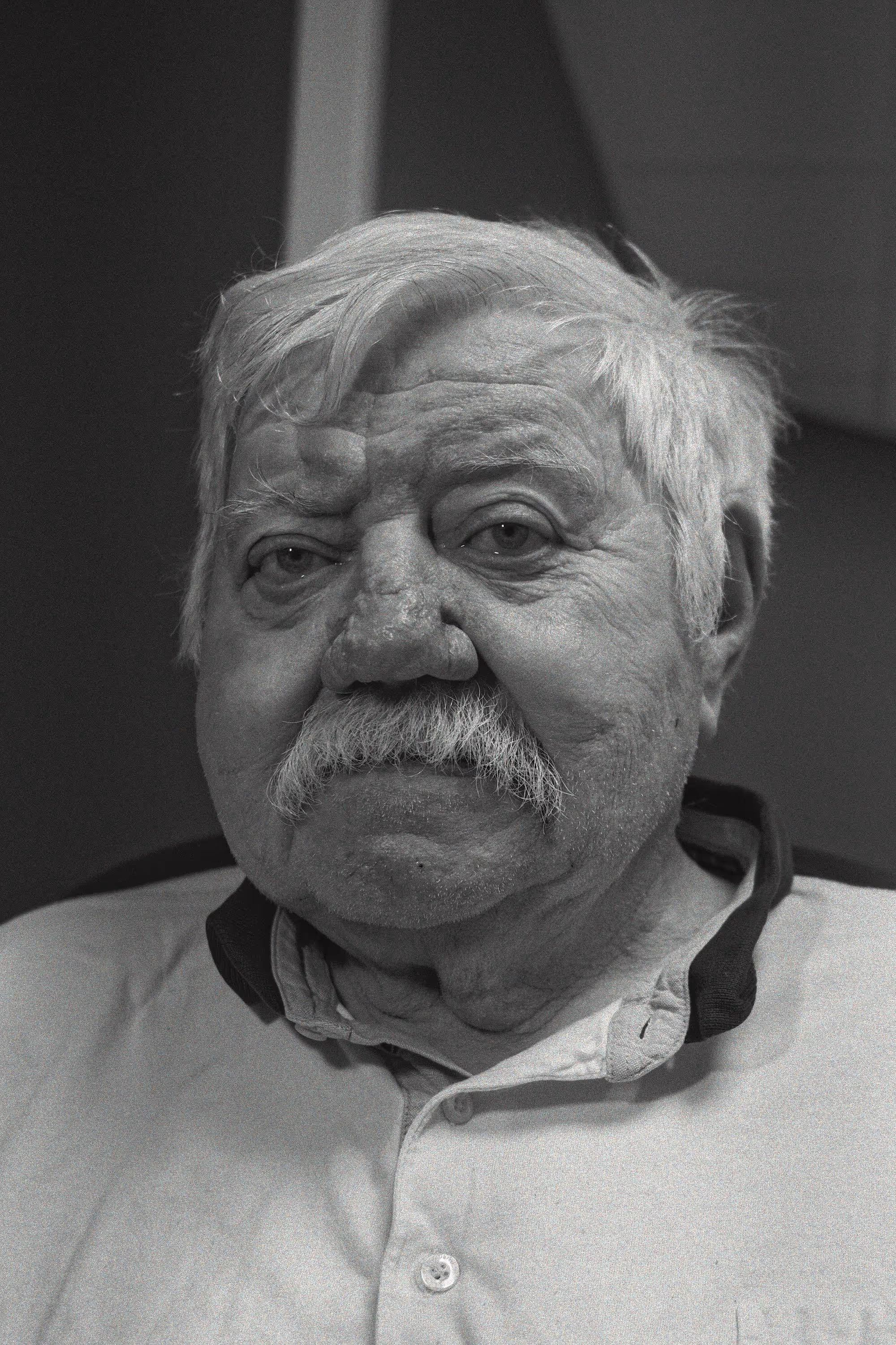 Portrait frontal d’un homme âgé moustachu, expression neutre, fond intérieur sombre.
