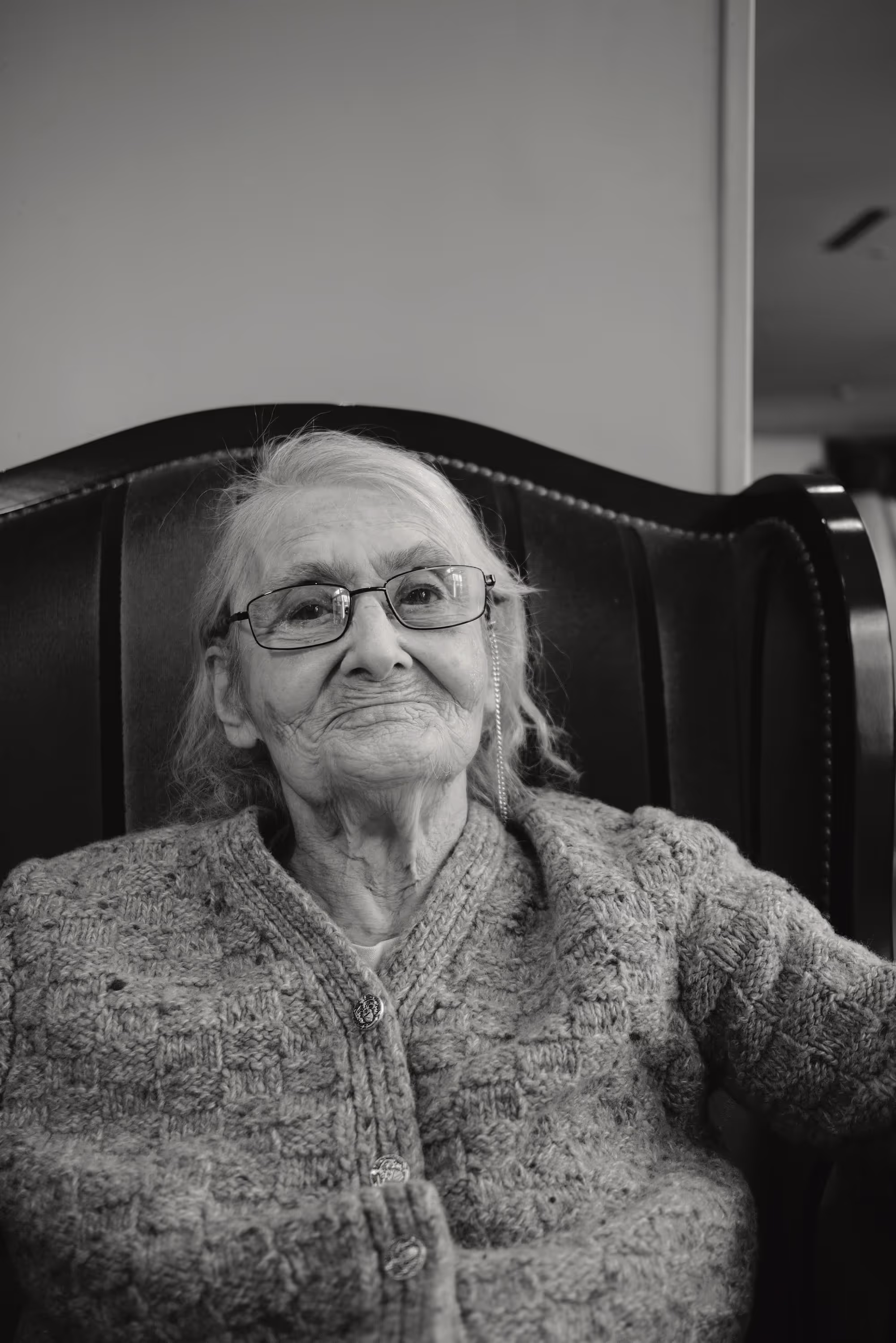 Femme âgée assise dans un fauteuil rembourré, portant des lunettes et un gilet tricoté, expression douce.