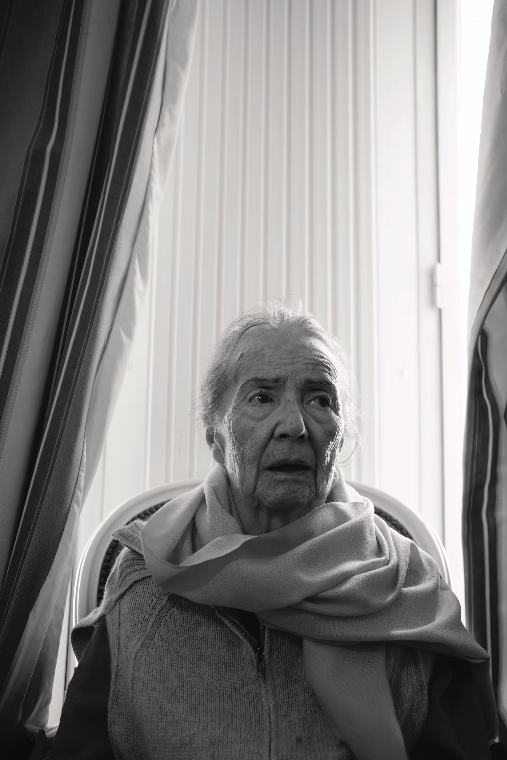 Femme âgée assise devant une fenêtre avec des rideaux, portant une écharpe épaisse, visage sérieux.