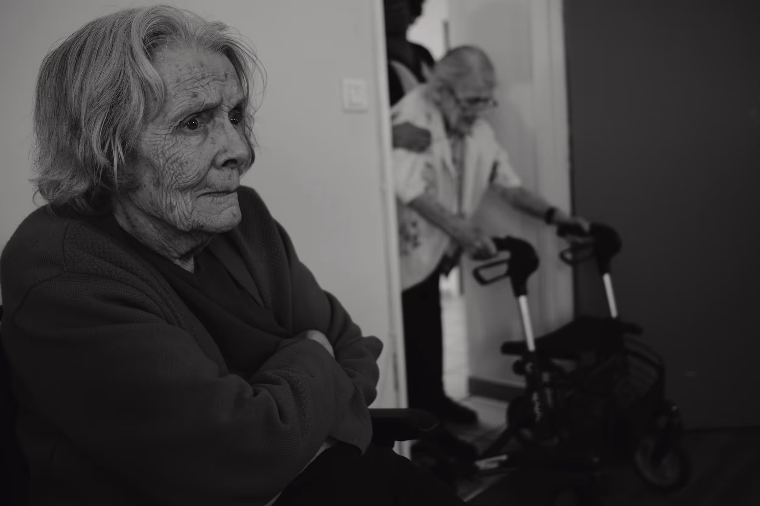 Femme âgée en gros plan de profil, assise en fauteuil roulant, une autre femme âgée entre dans la pièce en arrière-plan.