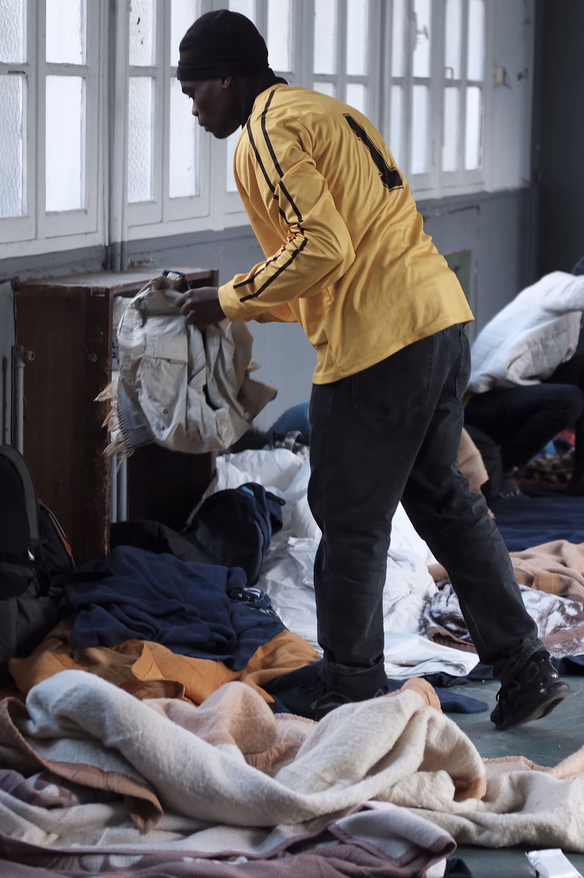 Un homme portant un maillot jaune ramasse ou plie des vêtements à l’intérieur d’un dortoir improvisé.