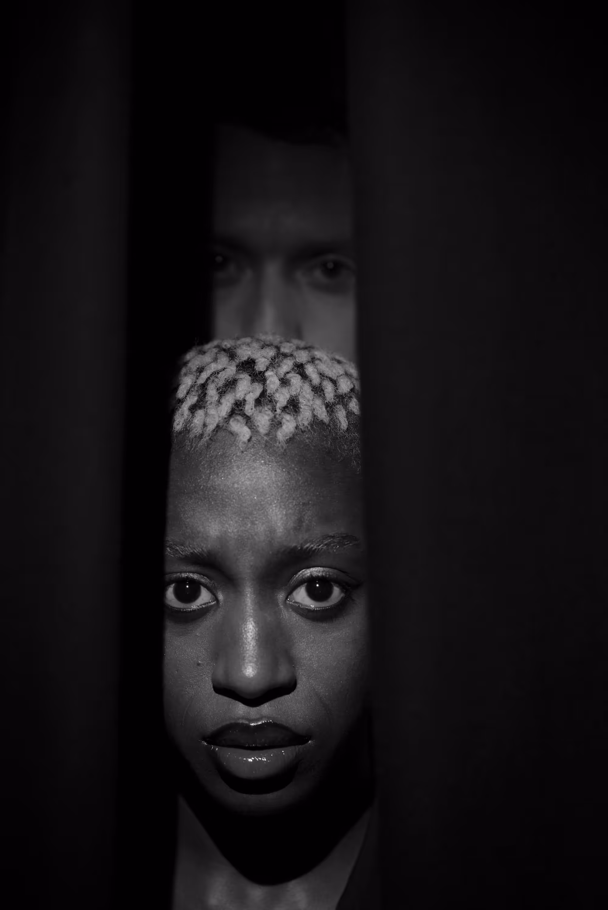 Portrait dramatique d’une femme regardant fixement à travers une ouverture entre deux rideaux sombres.