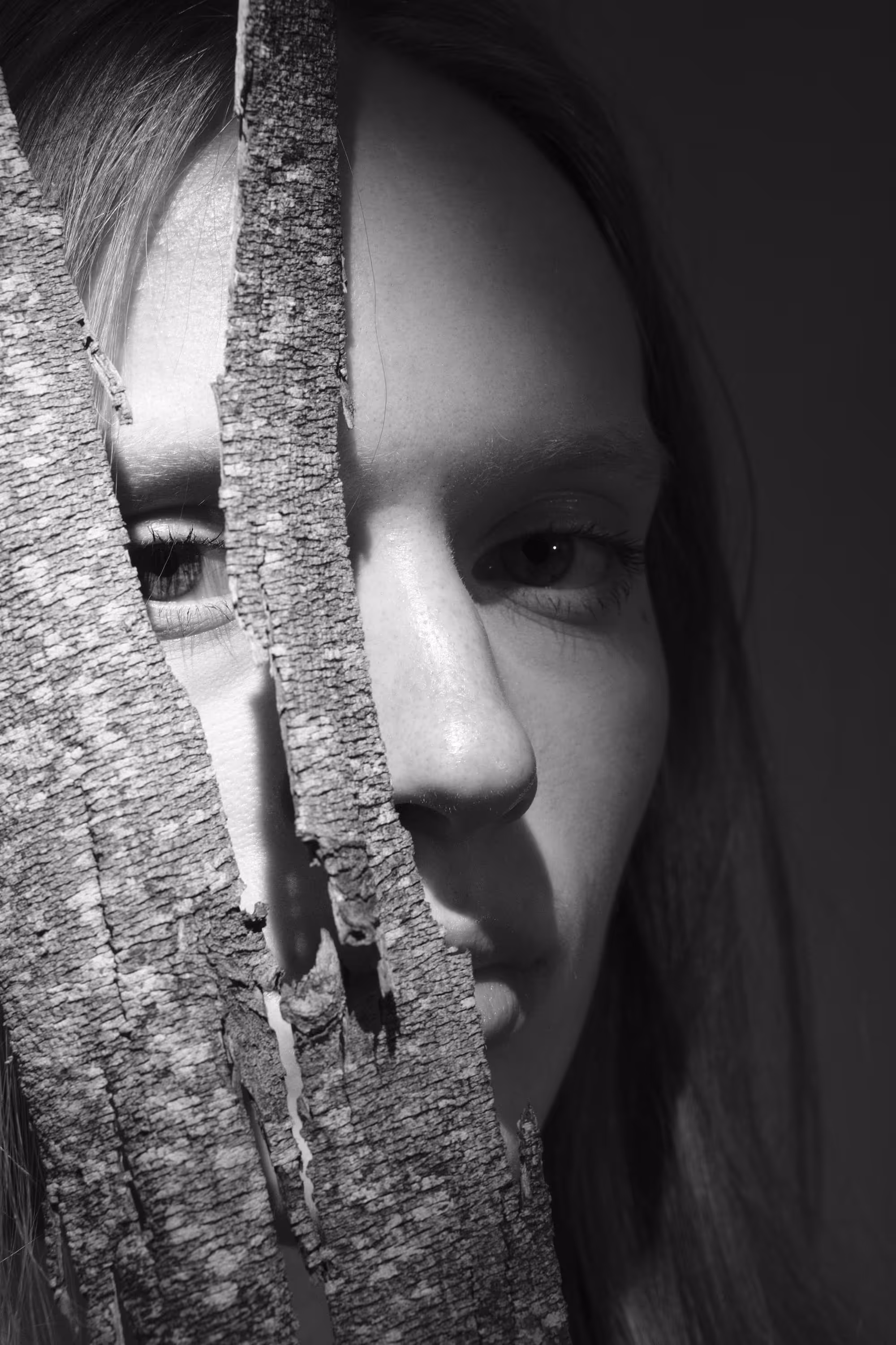 Portrait en noir et blanc d’une femme partiellement dissimulée derrière un morceau d’écorce, jouant sur la texture et la lumière.