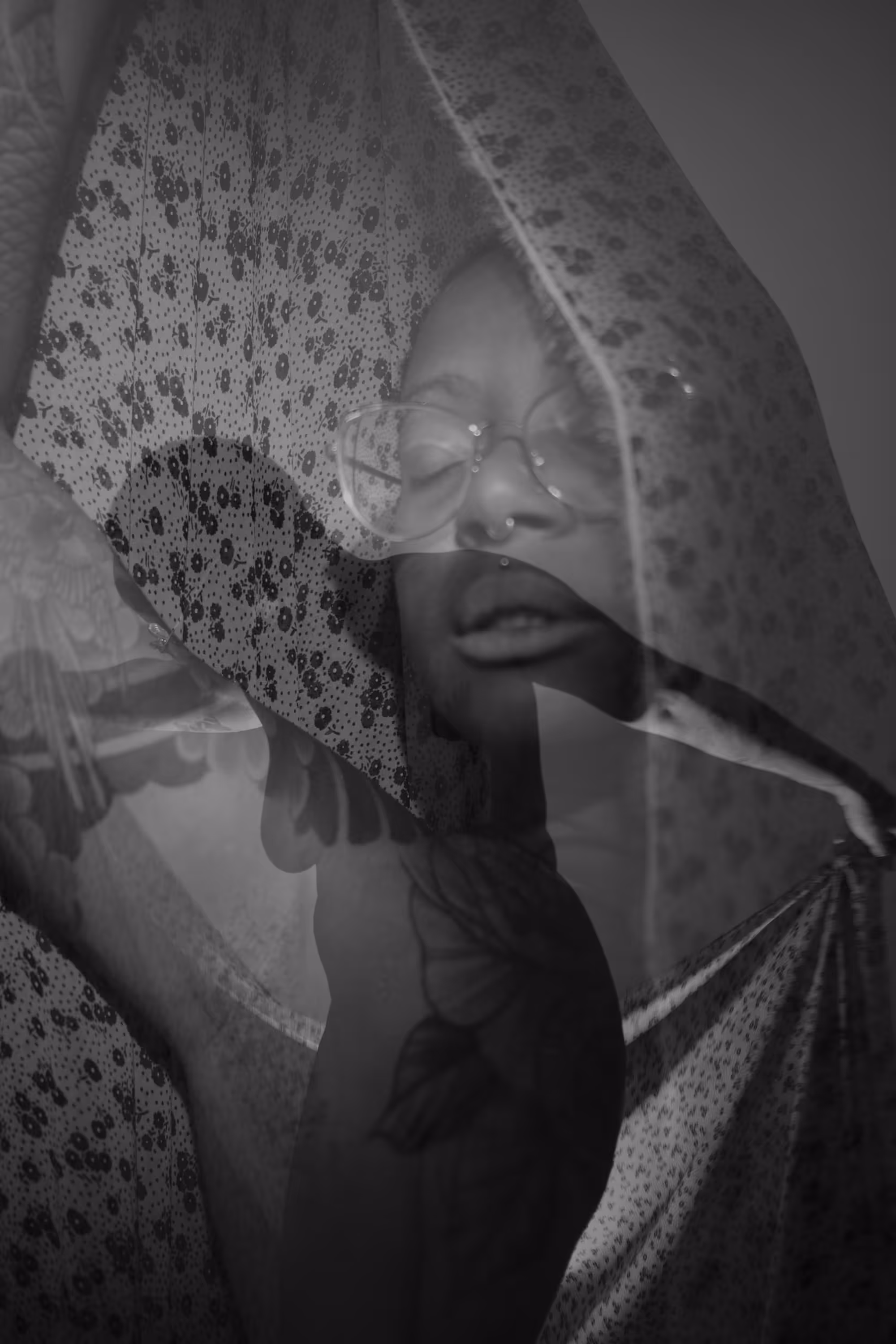Portrait poétique en double exposition d'une femme dansant avec des lunettes et un voile à motifs fleuris, yeux fermés, créant une sensation de mystère et de protection.