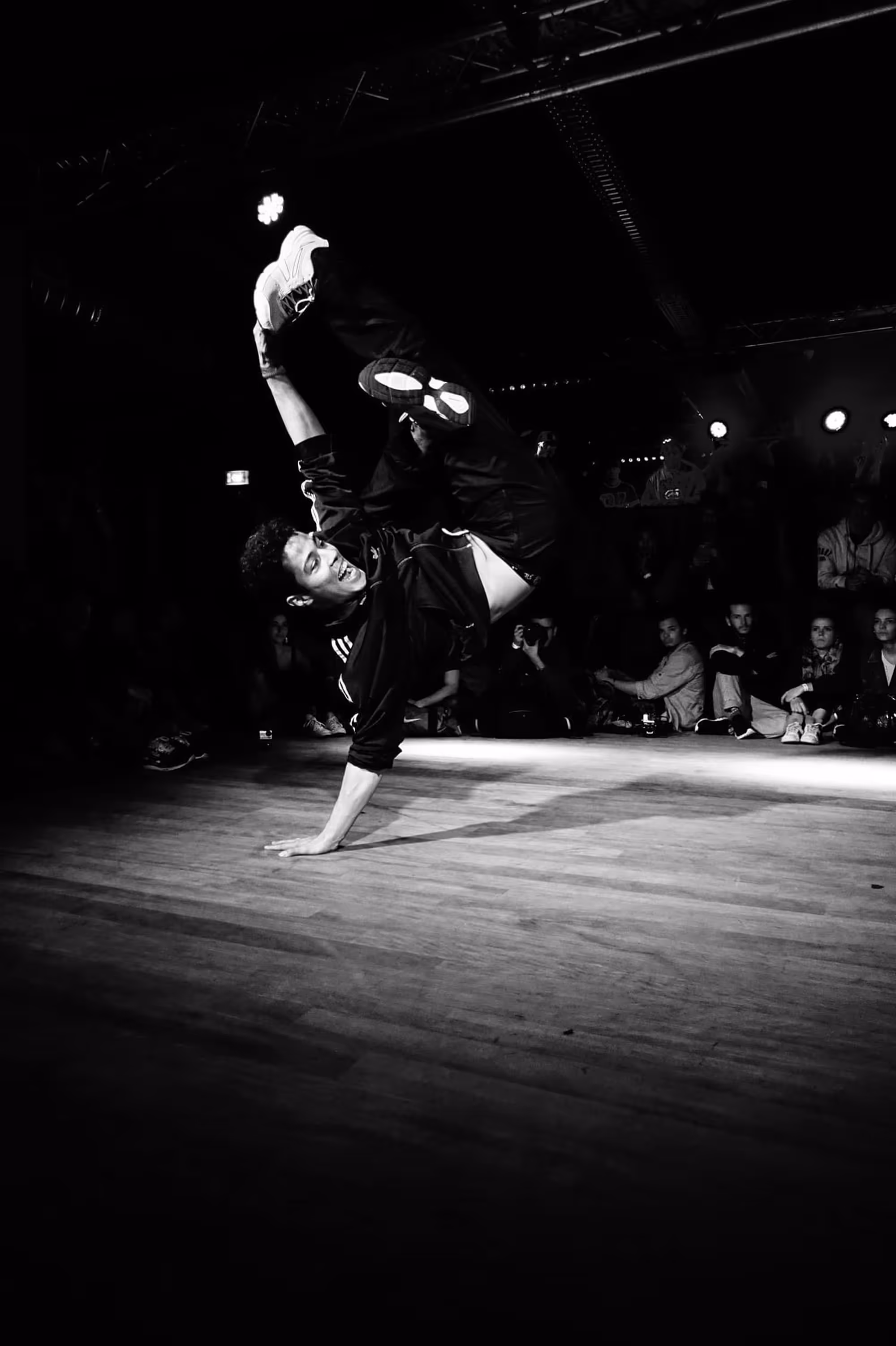 Danseur en équilibre sur une main, jambes pliées en l’air, éclairage de scène concentré.