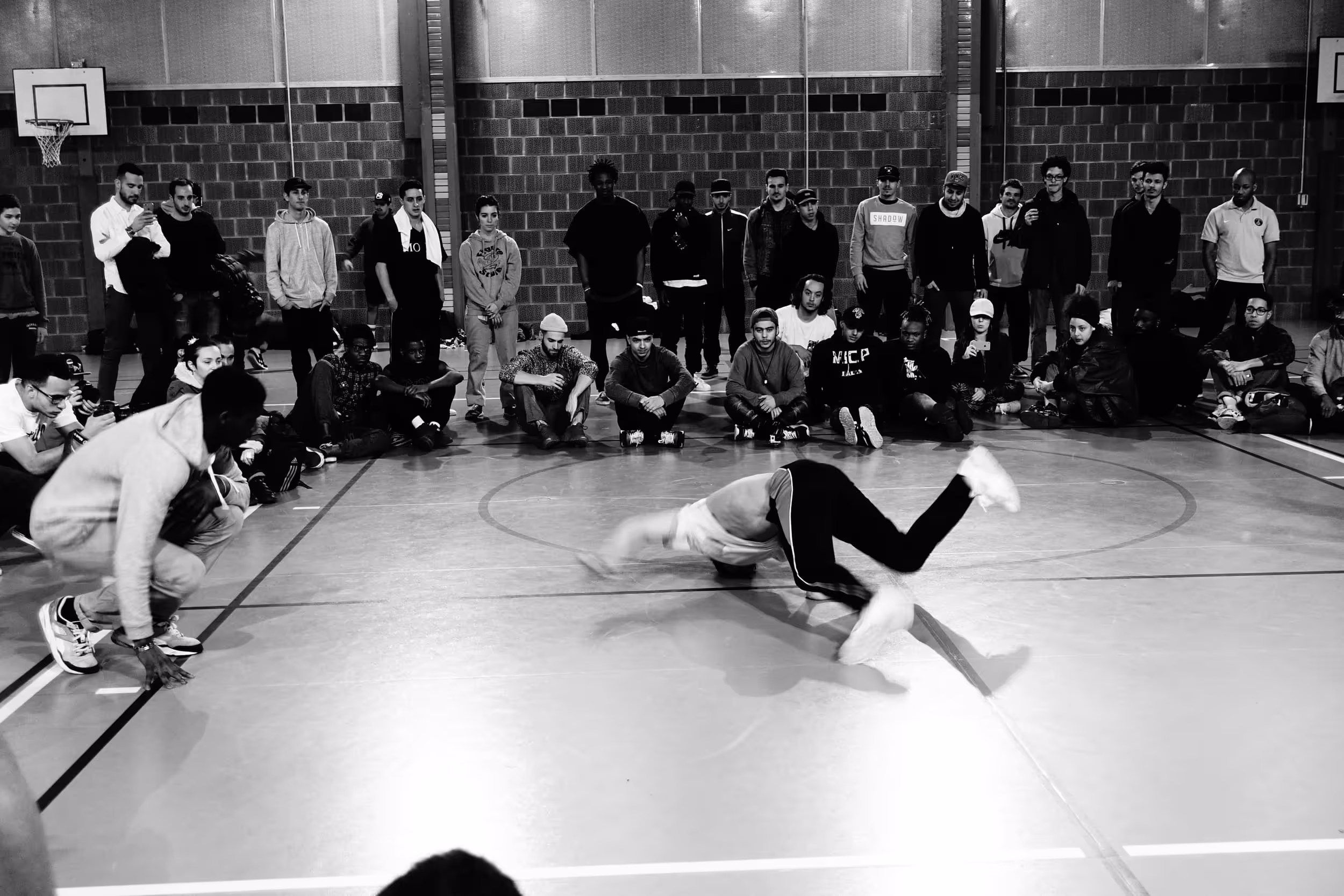 Compétition de danse dans un gymnase, avec un danseur flou en mouvement au centre du cercle formé par le public.