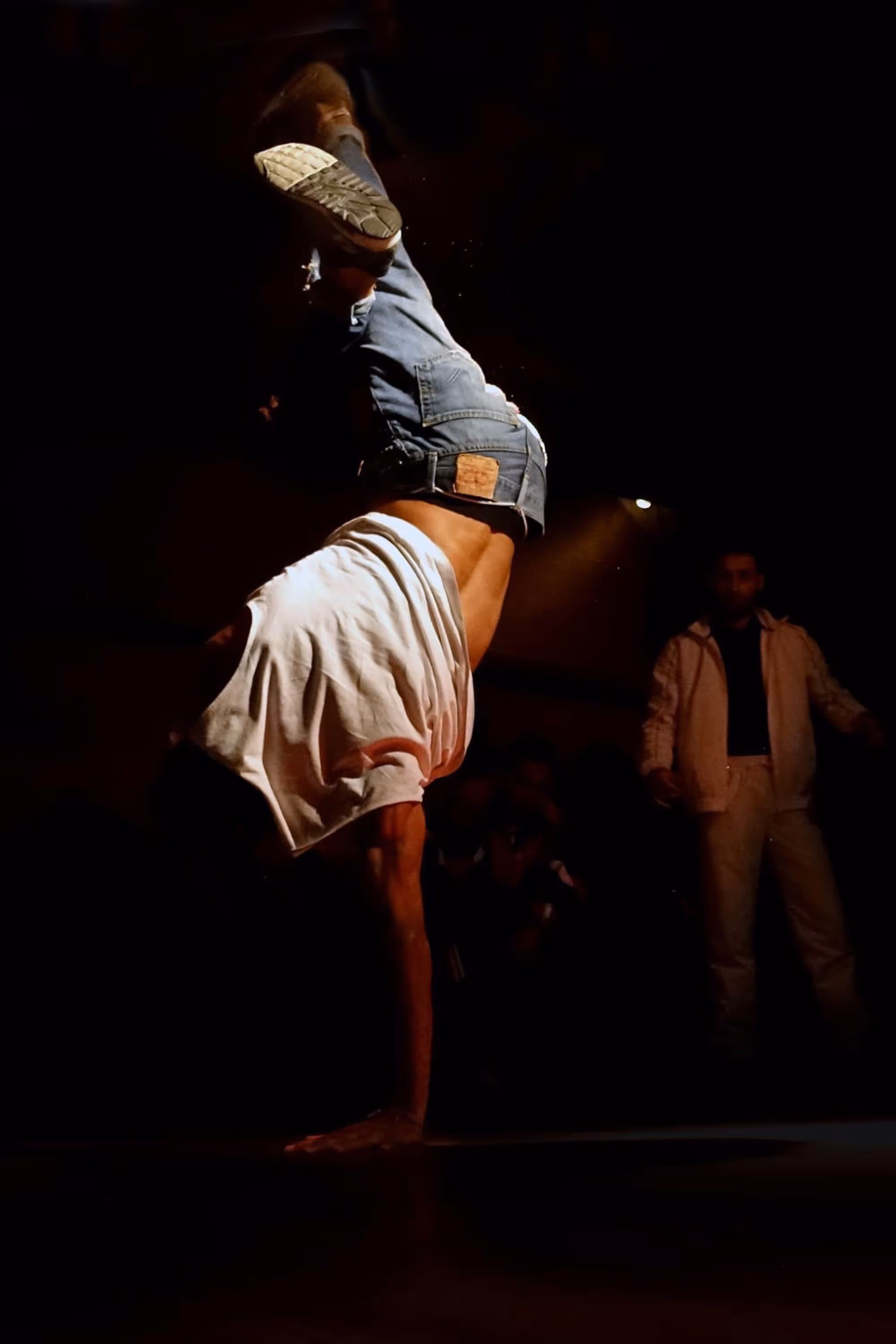 Danseur effectuant un équilibre sur une main sous un projecteur, portant un jean et un t-shirt blanc.