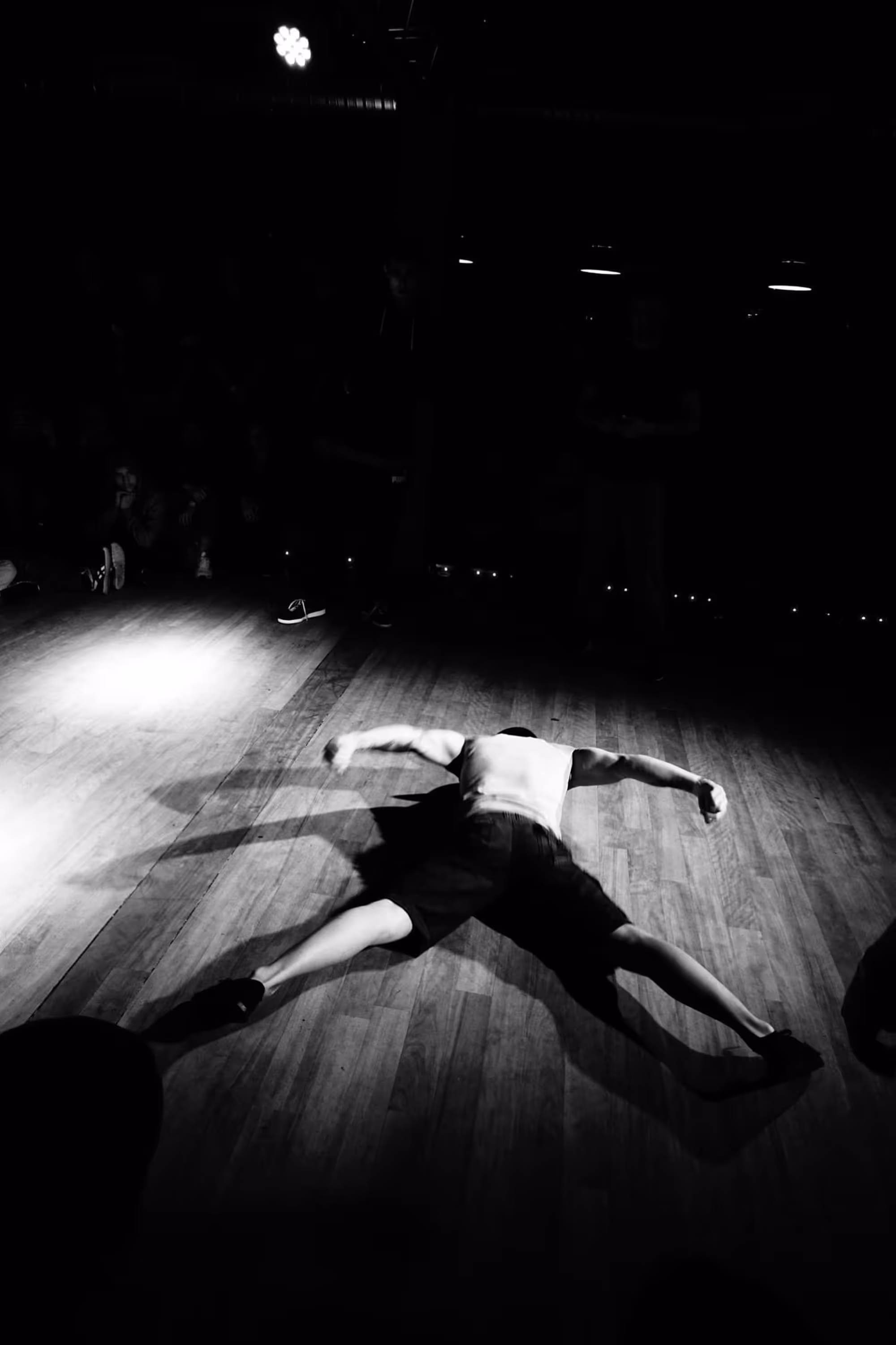 Danseur allongé en étoile sur le sol en bois, sous un projecteur intense, ambiance dramatique.