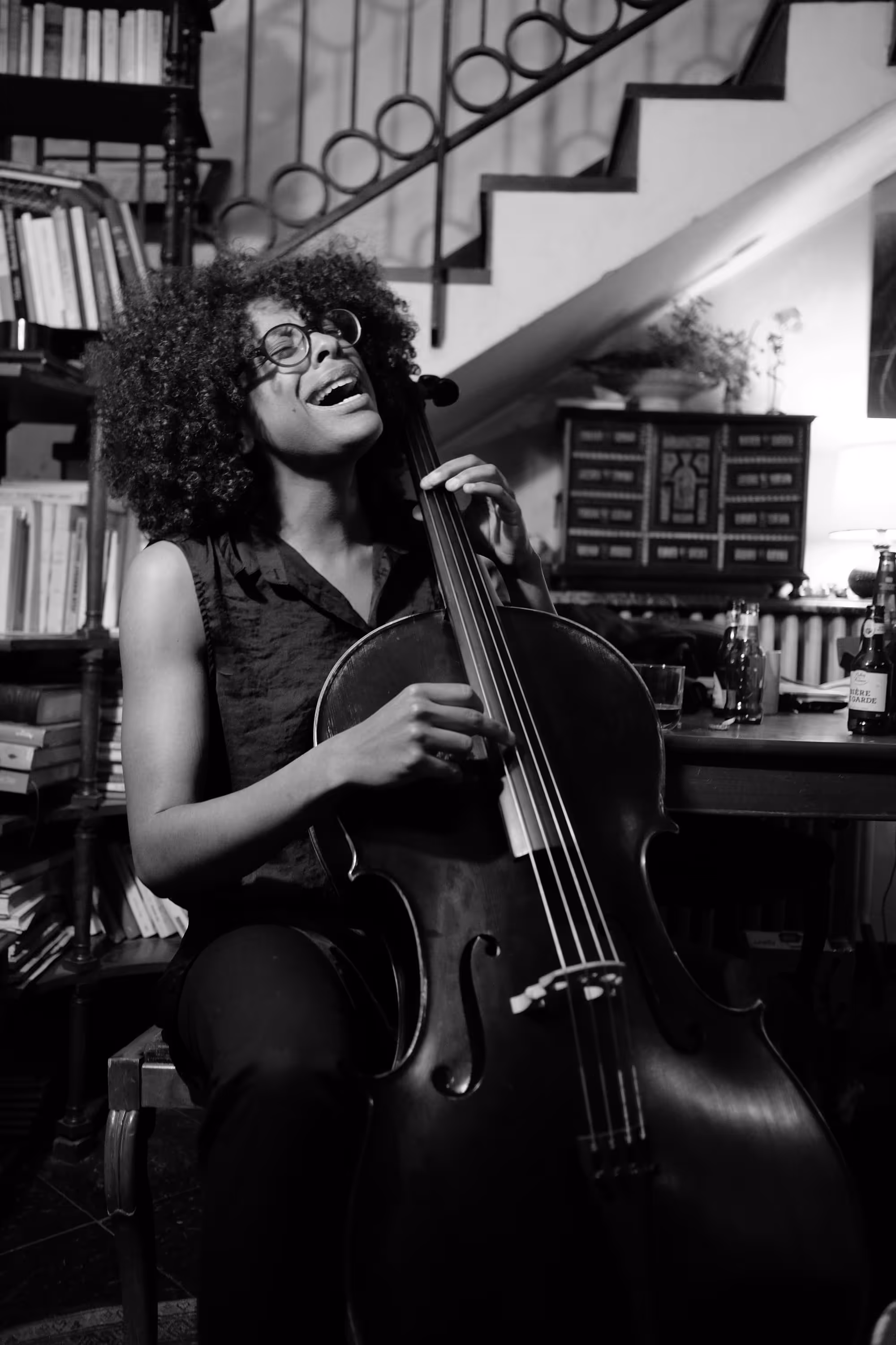 Violoncelliste chantant en jouant, dans un intérieur avec bibliothèque et escalier.