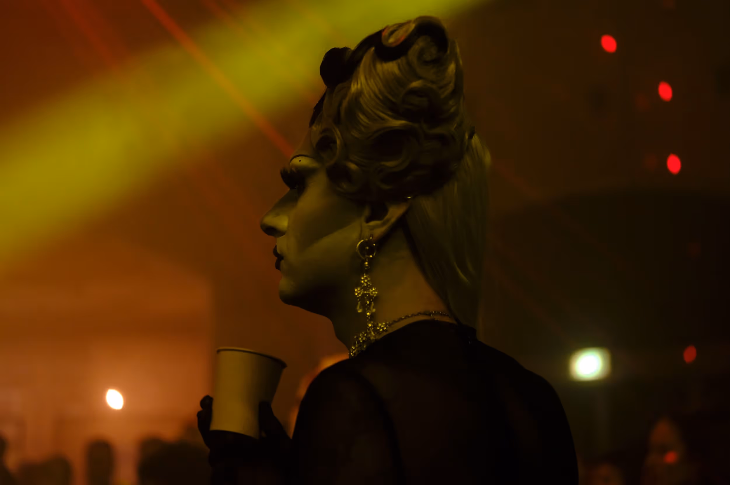 Personne en drag dans un club sombre, tenant un gobelet, éclairée par des lumières colorées.
