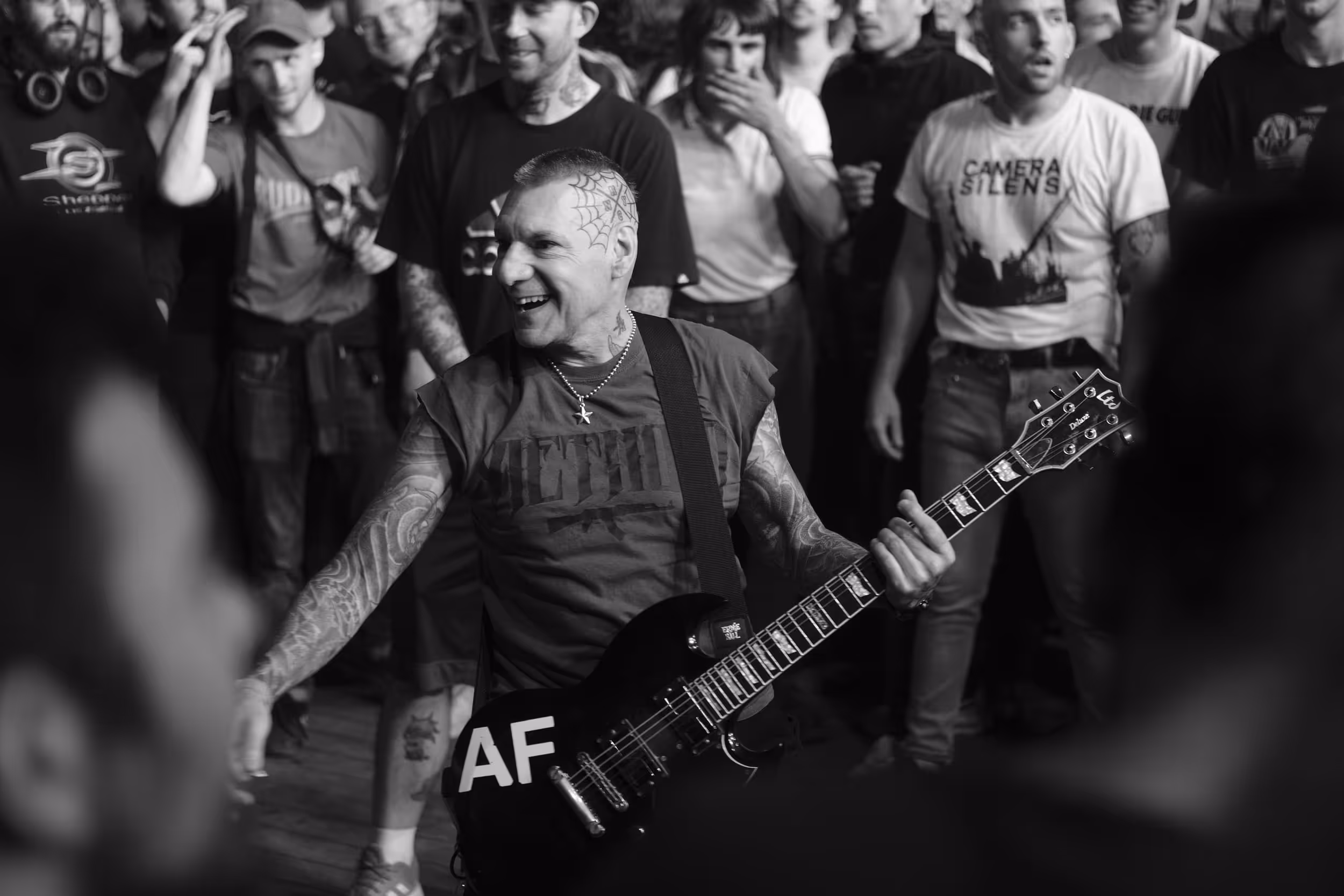 Guitariste tatoué souriant au centre d’une foule enthousiaste lors d’un concert punk.