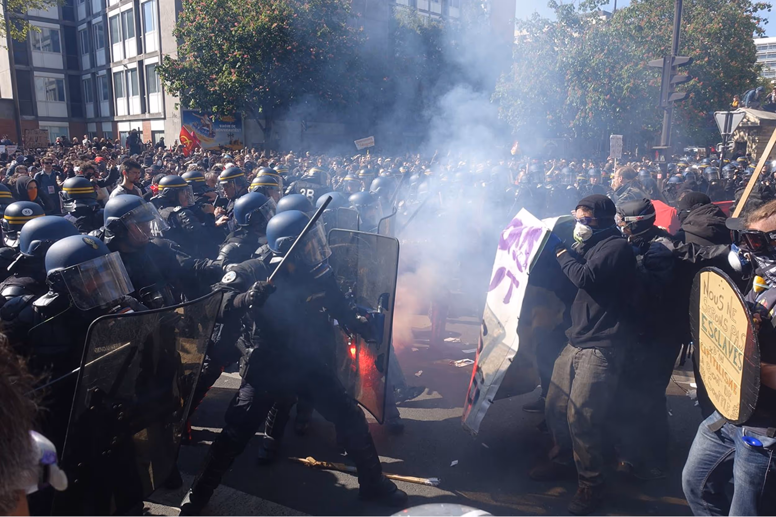 Affrontement entre policiers en tenue anti-émeute avec boucliers et manifestants masqués tenant des pancartes, dans une rue enfumée.
