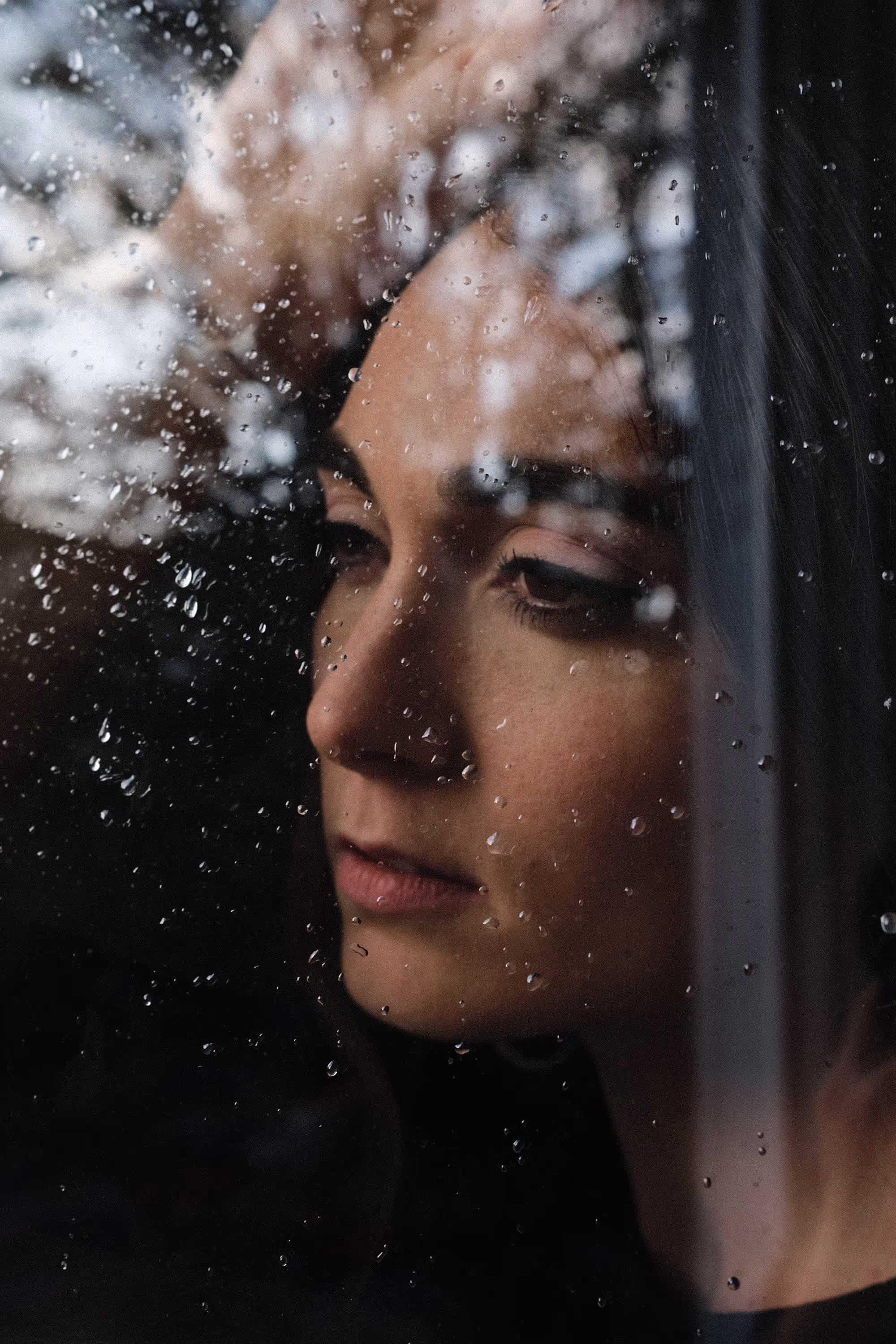 Portrait d'une femme pensive regardant à travers une vitre couverte de gouttes de pluie.