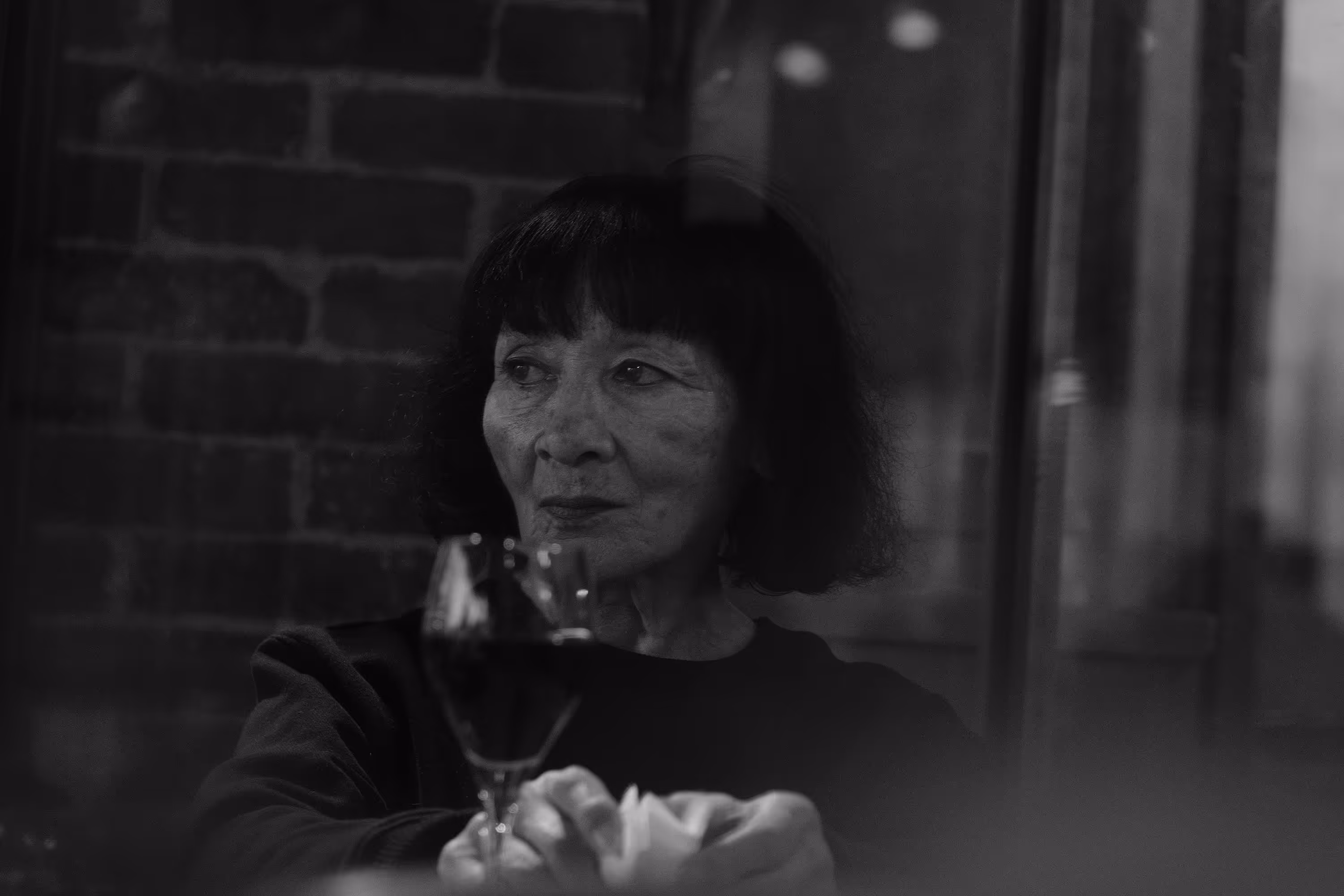 Femme asiatique aux cheveux courts regardant sur le côté à travers une fenêtre avec un verre de vin rouge devant elle.
