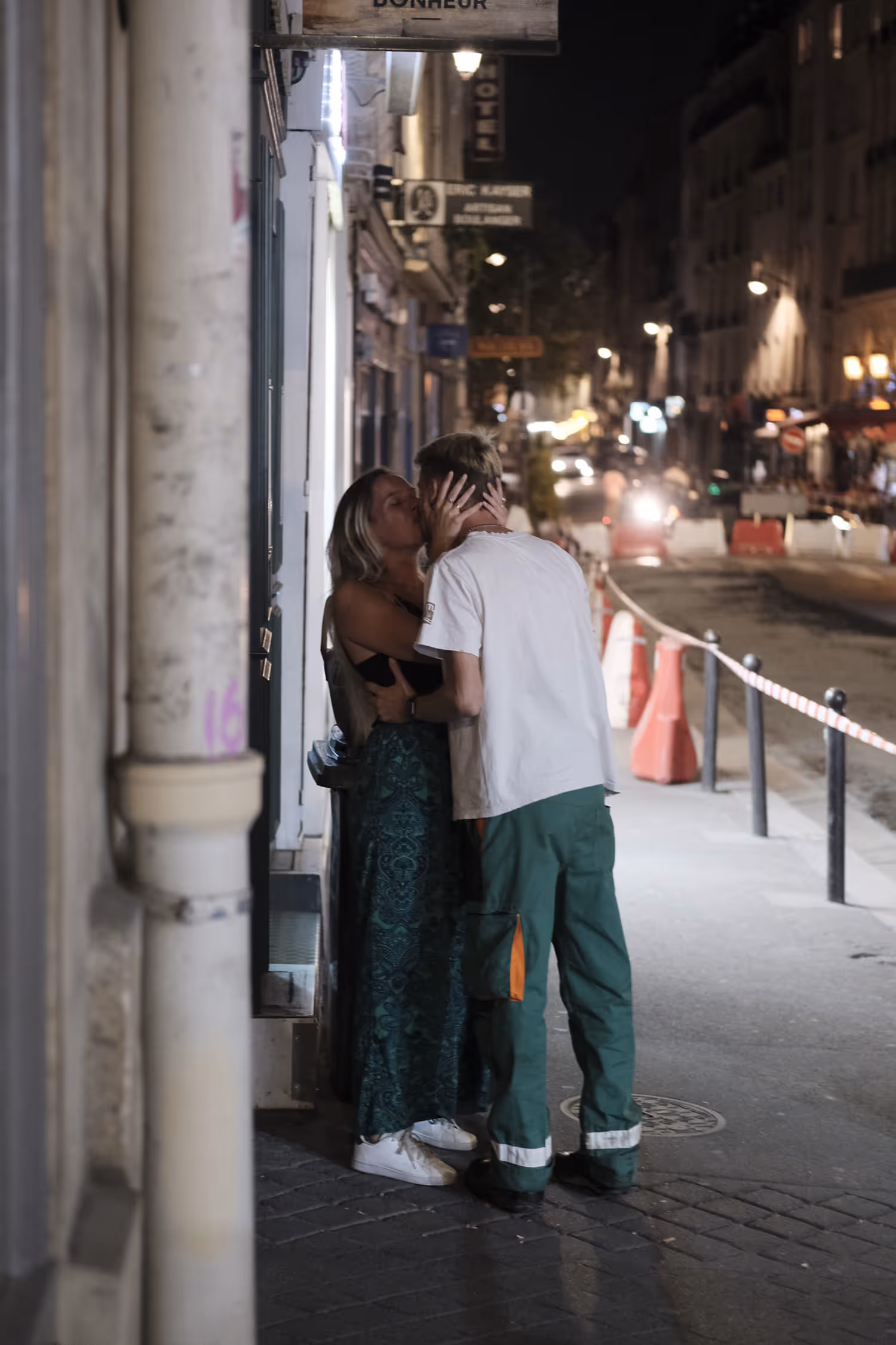 Un couple s'embrassant tendrement sur un trottoir de rue la nuit, avec des lumières urbaines en arrière-plan.