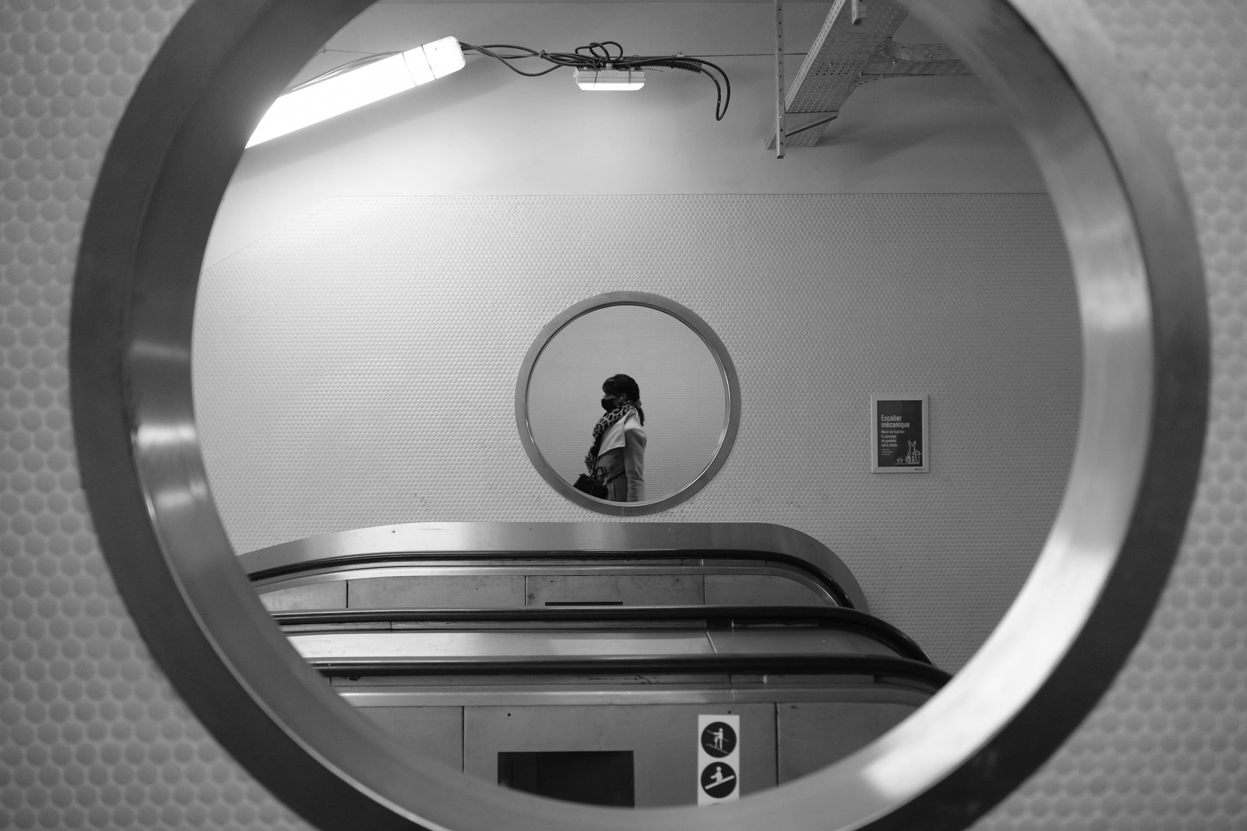 Personne masquée marchant dans une station de métro, vue à travers un grand cercle métallique avec un petit miroir rond encadrant la personne.