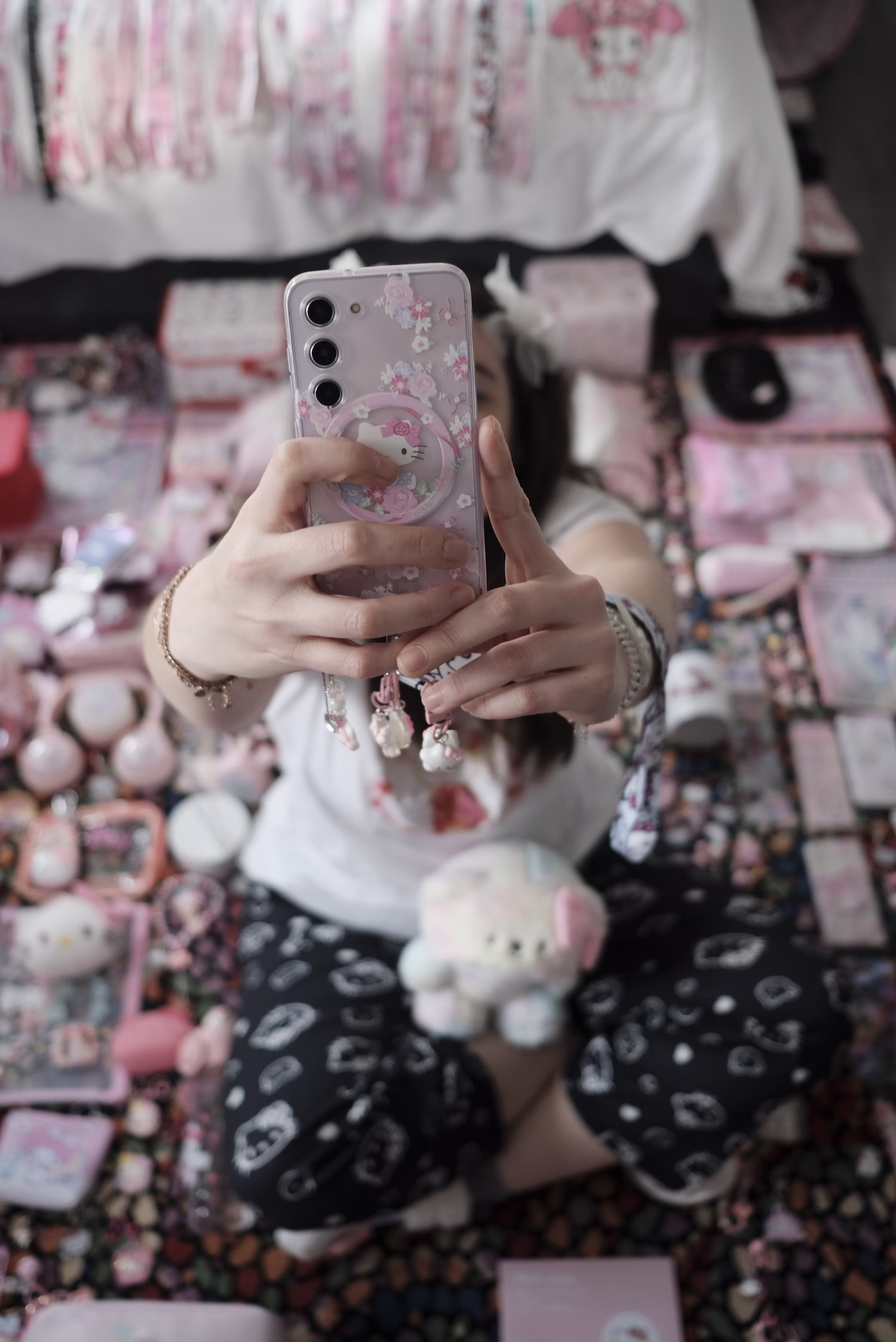 Person assise par terre prenant un selfie avec un téléphone rouge à motifs floraux, entourée d'objets décoratifs et accessoires roses disposés autour d'elle, y compris une peluche rose et des articles assortis sur un tapis coloré.