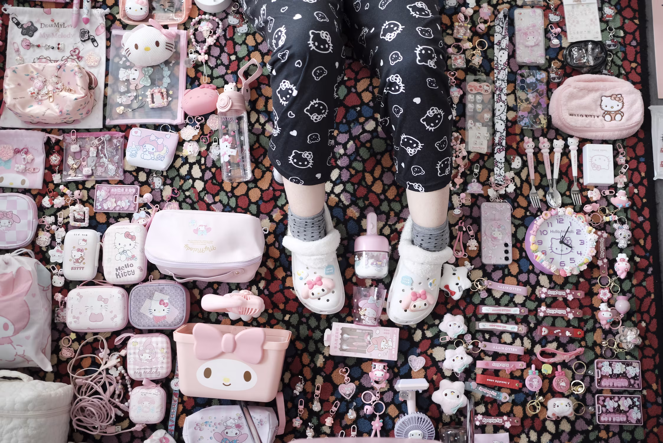Une personne portant un pantalon et des chaussons Hello Kitty, entourée d'articles roses Hello Kitty et My Melody, sur un tapis coloré.