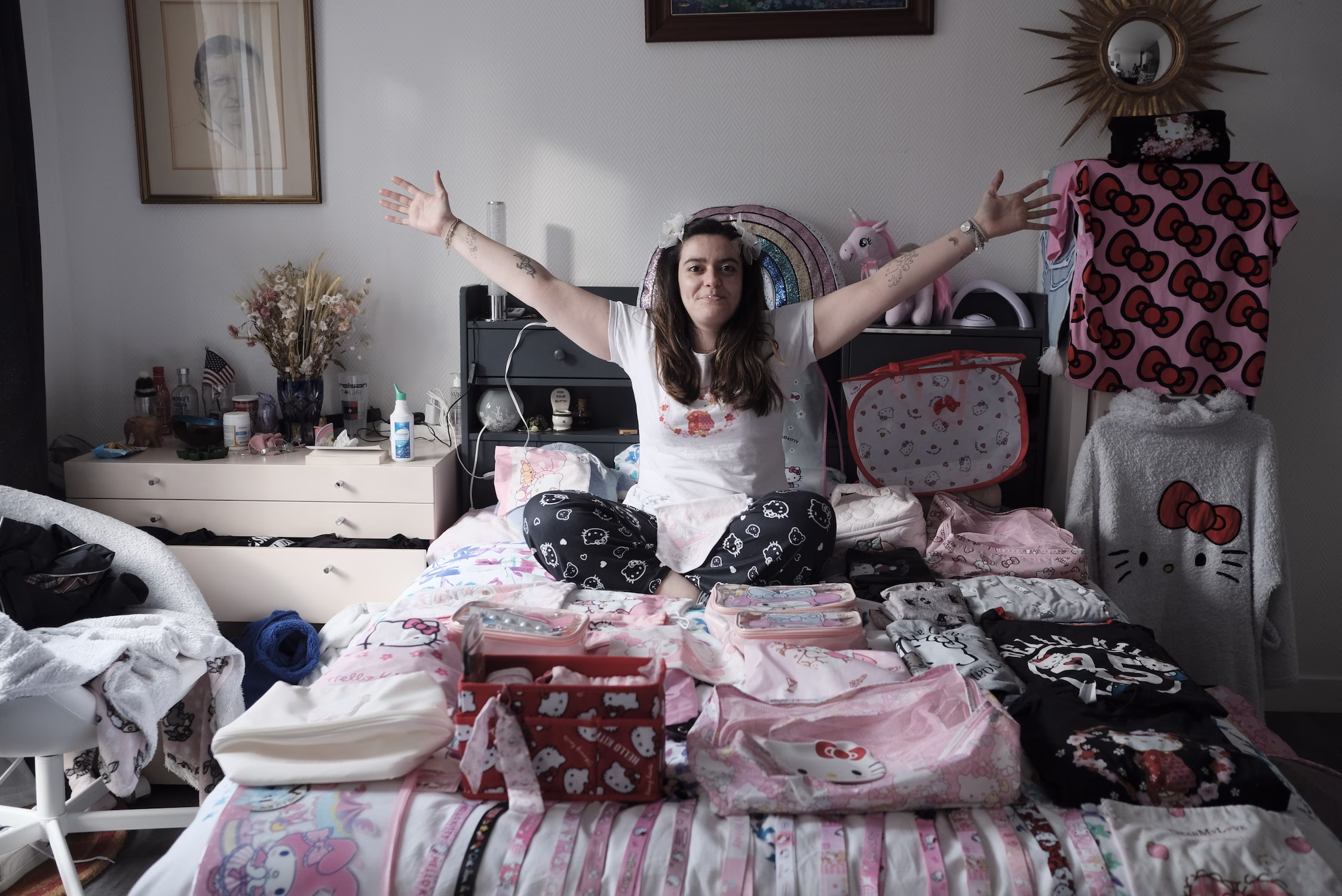 Une femme assise sur un lit d’articles Hello Kitty, bras écartés, dans une chambre décorée.