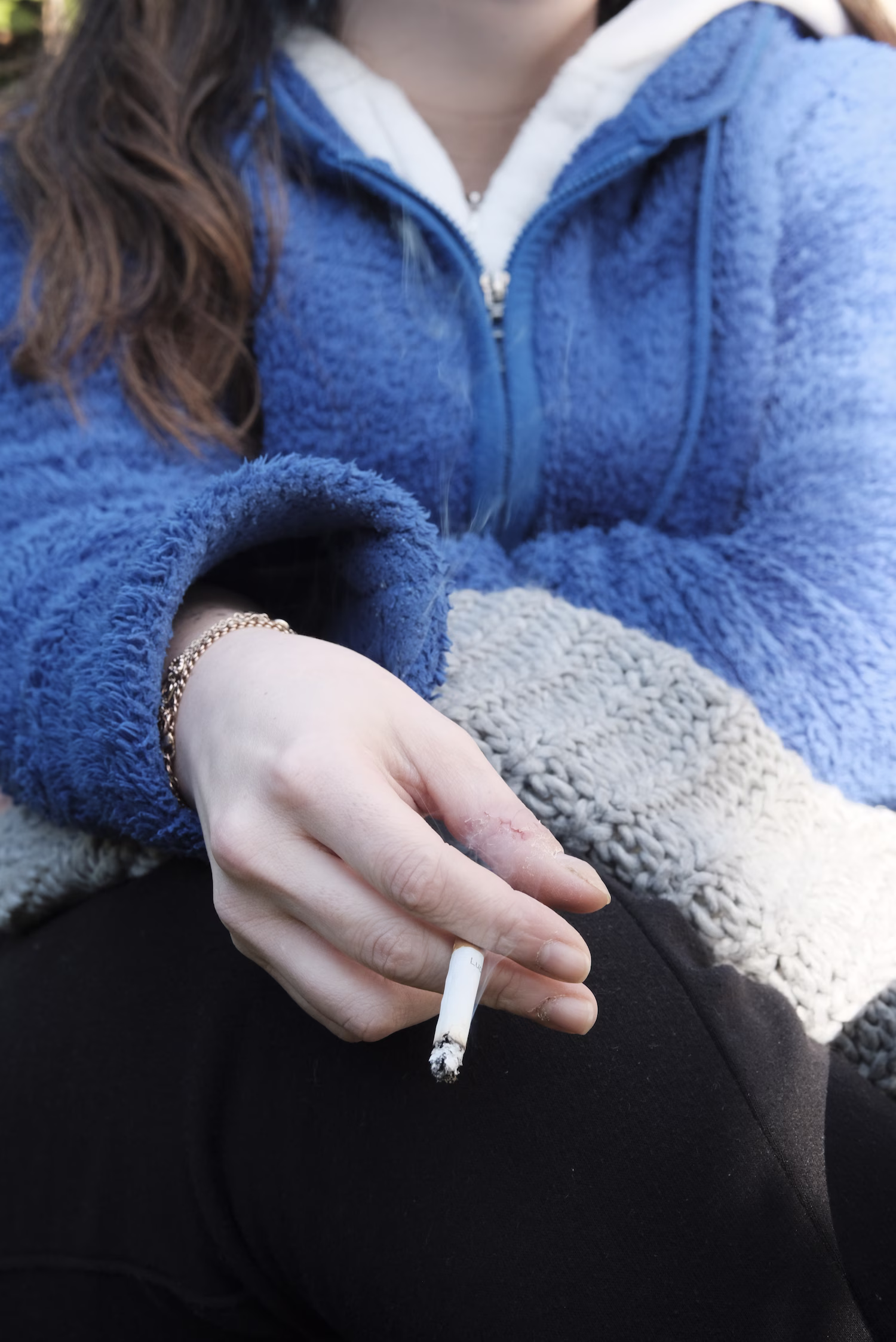 Une main tenant une cigarette allumée, vêtue d'une veste polaire bleue à manches longues.