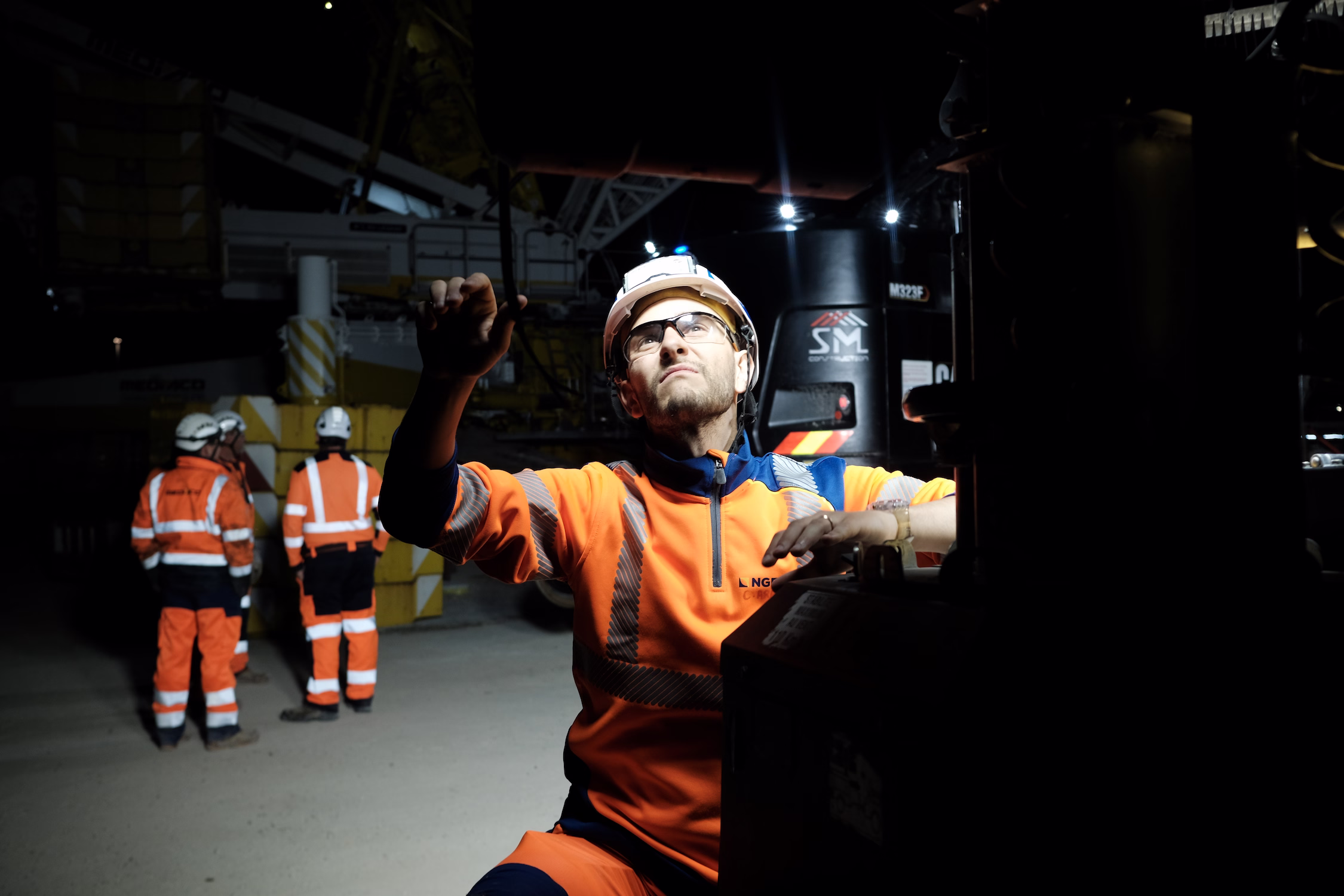 Ouvrier en tenue orange réfléchissante et casque inspectant une machine dans une usine la nuit.