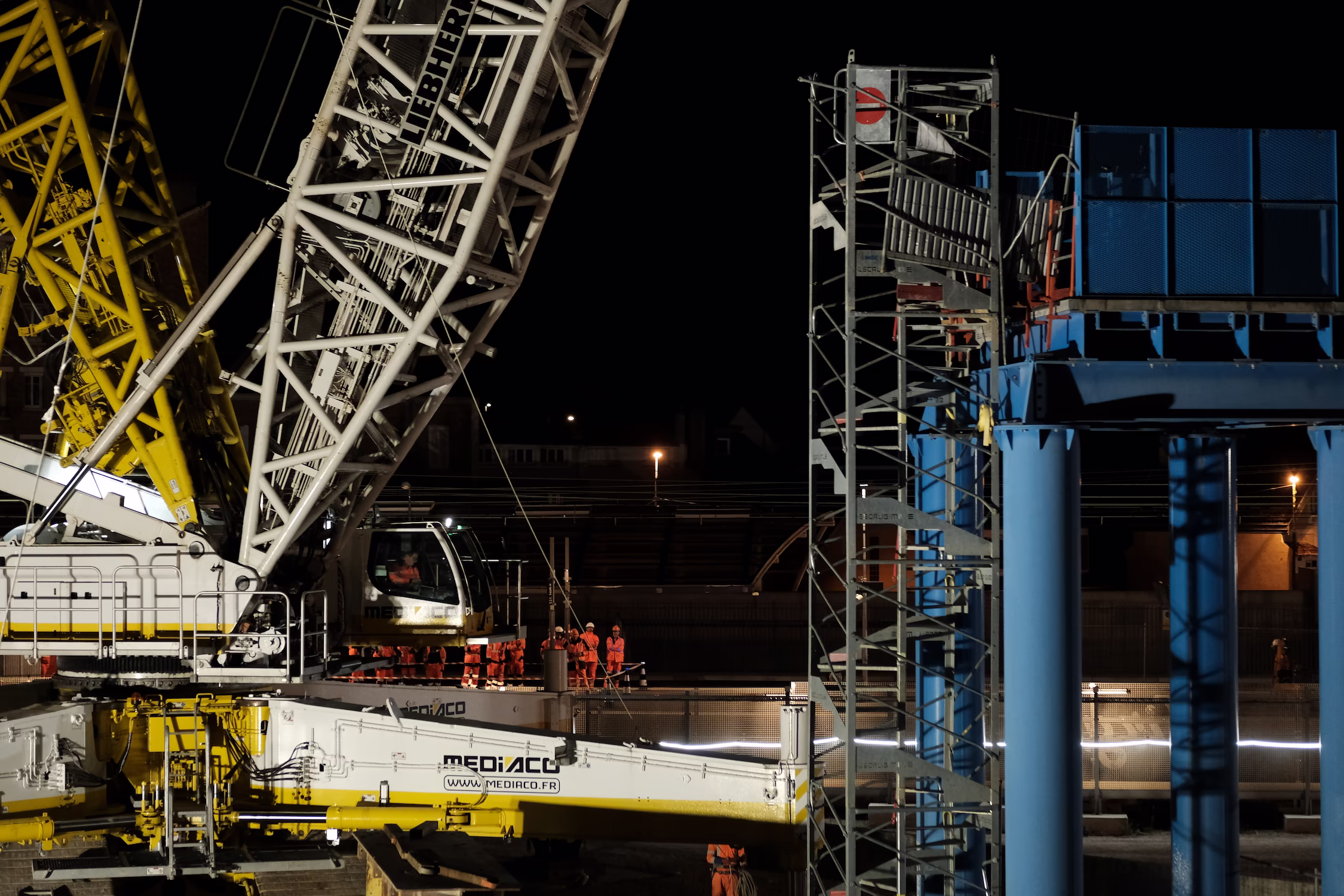 Grue et structure de chantier bleu éclairées la nuit avec ouvriers en tenue orange.