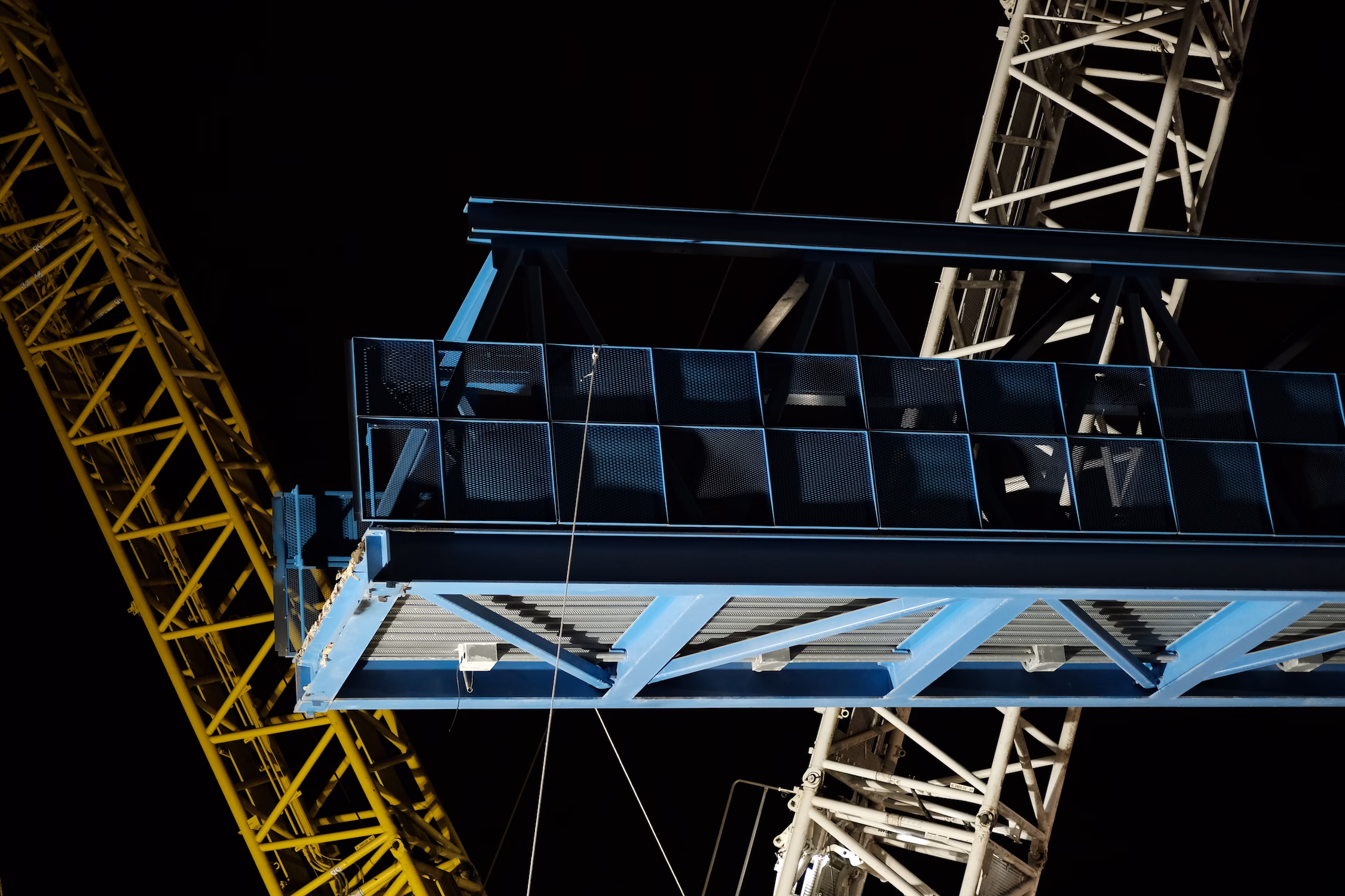 Passerelle métallique bleue et grues de construction jaune et blanche la nuit.