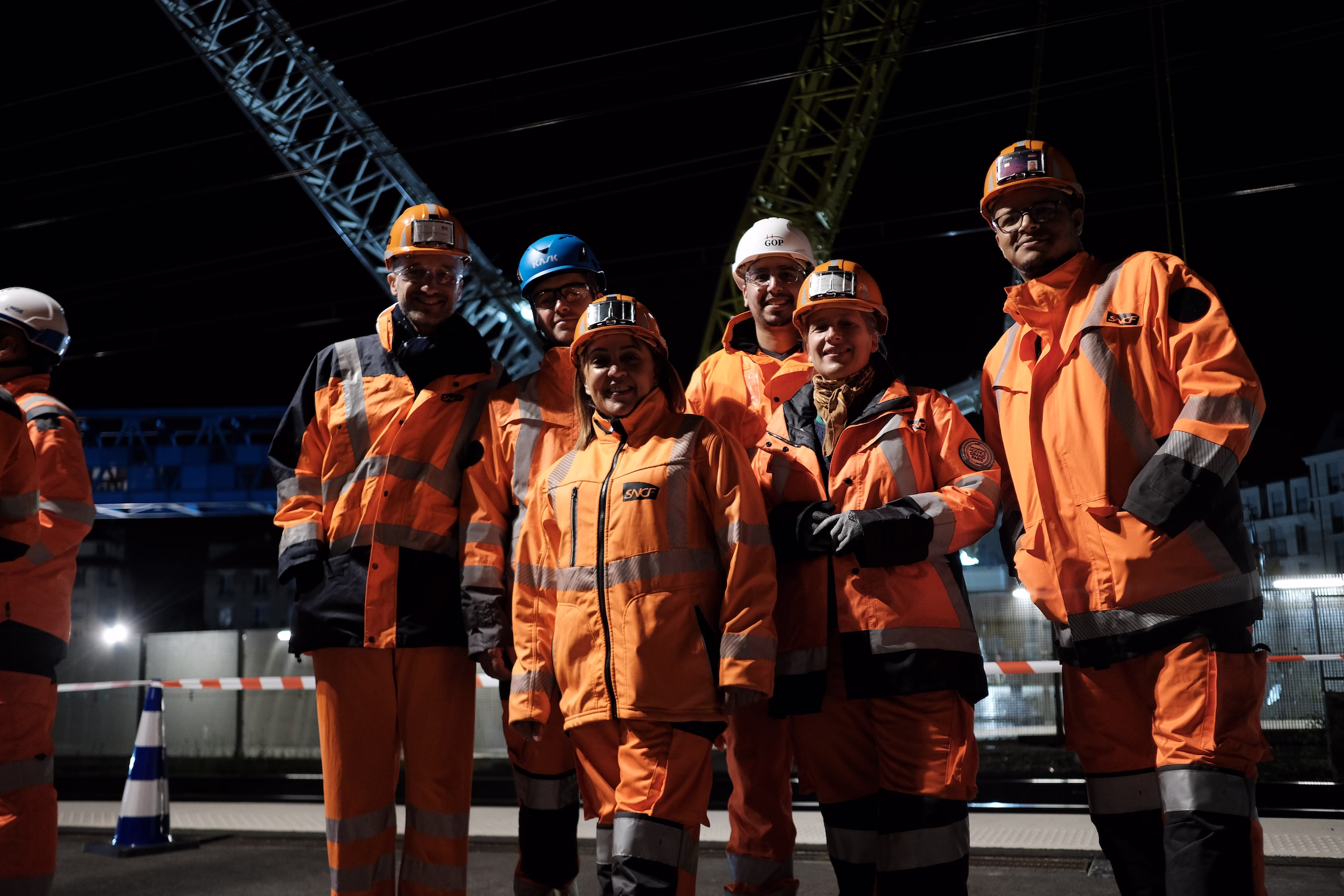 Groupe d’agents SNCF en tenues de sécurité orange posant de nuit sur un chantier ferroviaire.