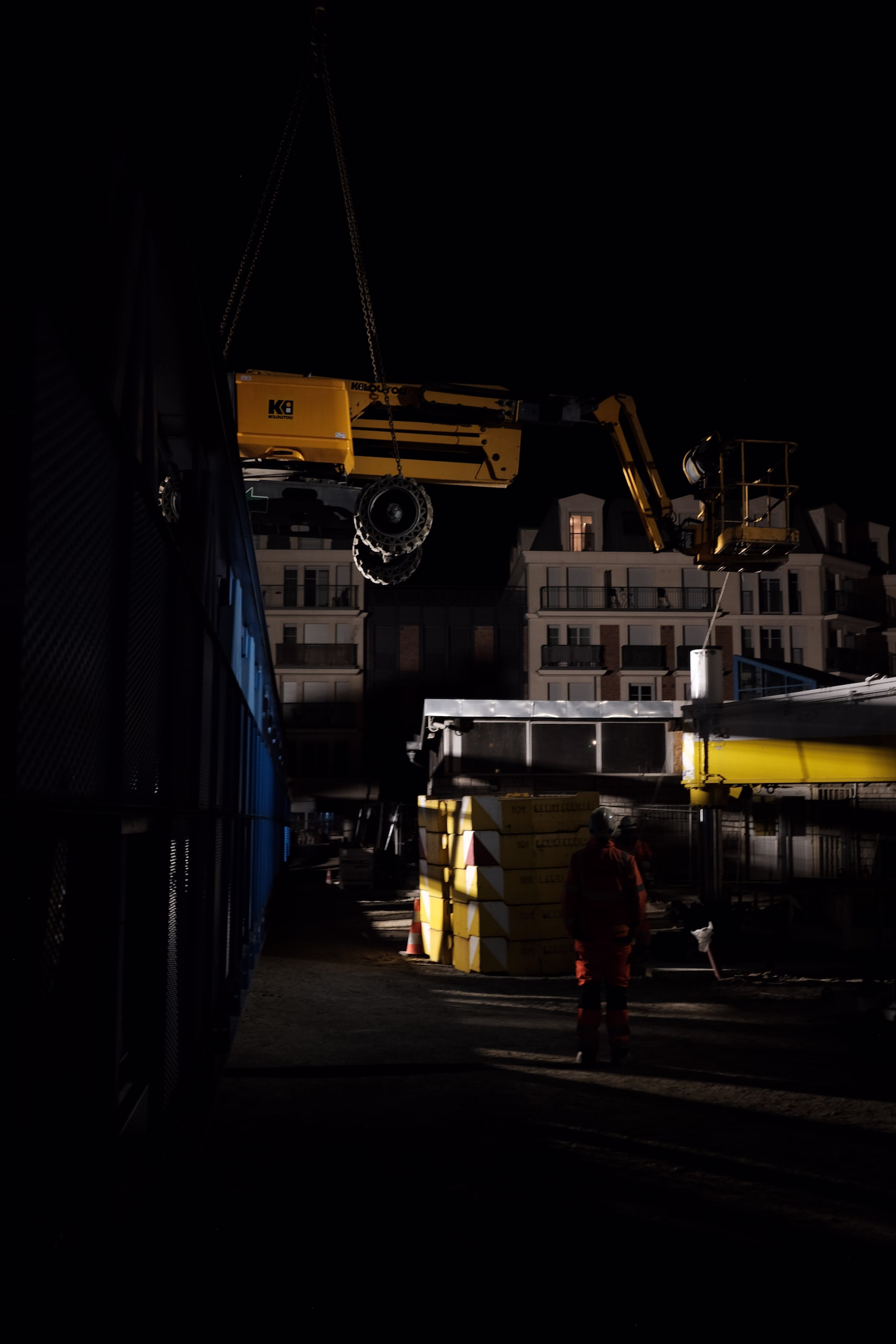 Engin de chantier jaune suspendu au-dessus d'un site avec un ouvrier en tenue orange la nuit.