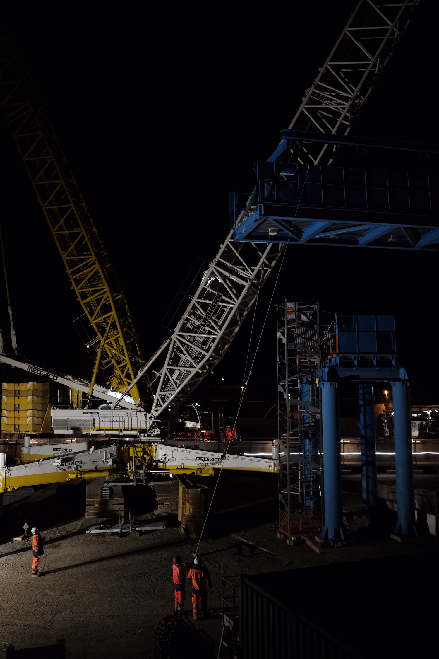 Grue de chantier jaune et blanche et travailleurs en orange sur un site de construction de nuit.