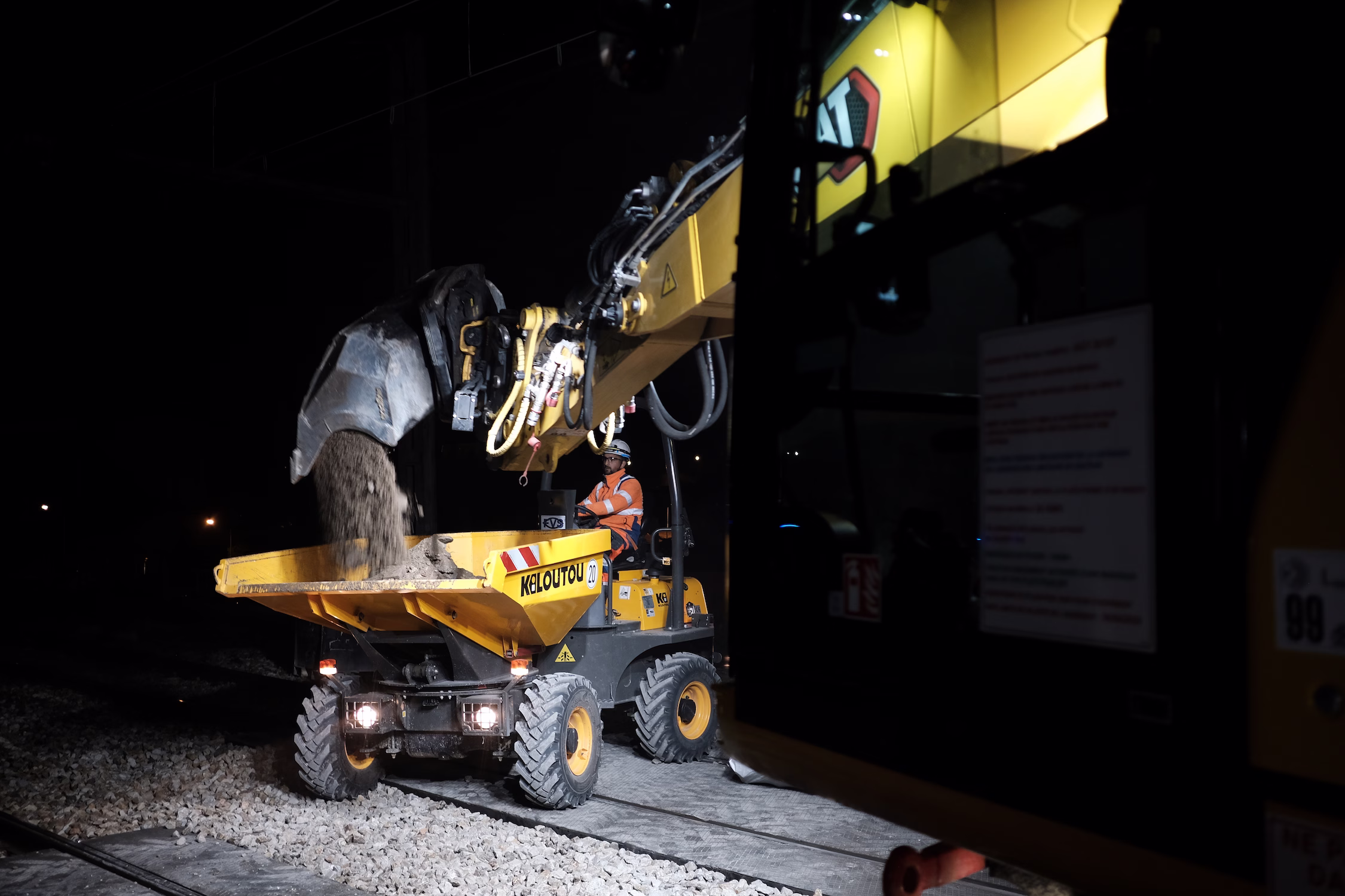 Un ouvrier en tenue orange conduit un mini-dumper jaune la nuit sur des gravats de chantier.