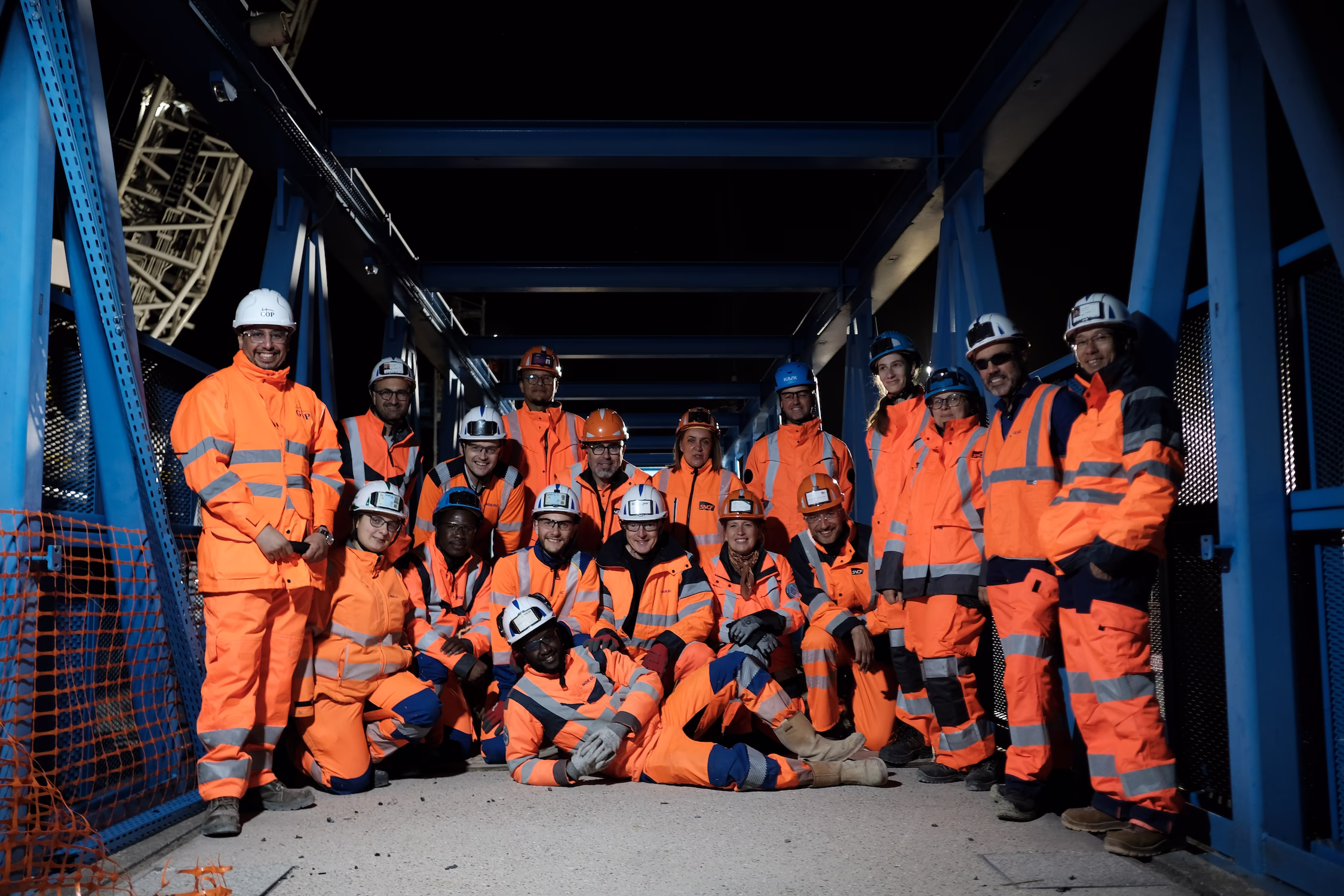 Groupe de travailleurs en tenue de sécurité orange avec casques posant dans une structure métallique bleue la nuit.