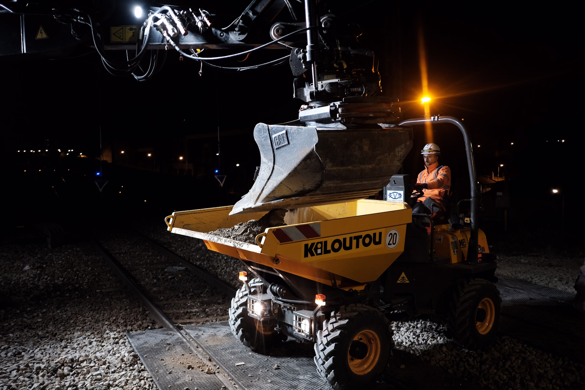 Chauffeur de camion-benne jaune Kiloutou chargé de gravats la nuit sur un chantier ferroviaire.