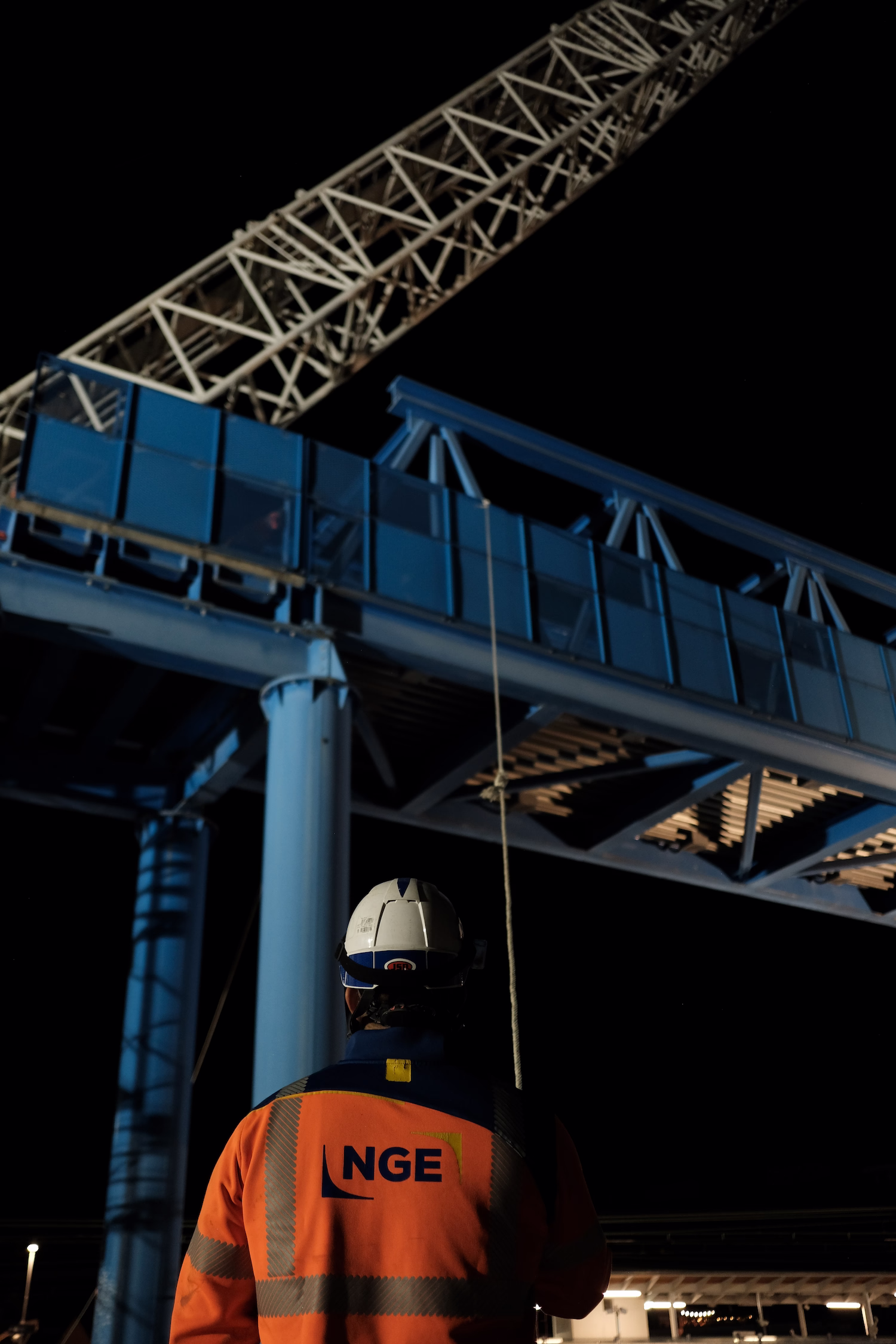 Ouvrier portant un casque et une veste NGE observe une passerelle bleue la nuit.