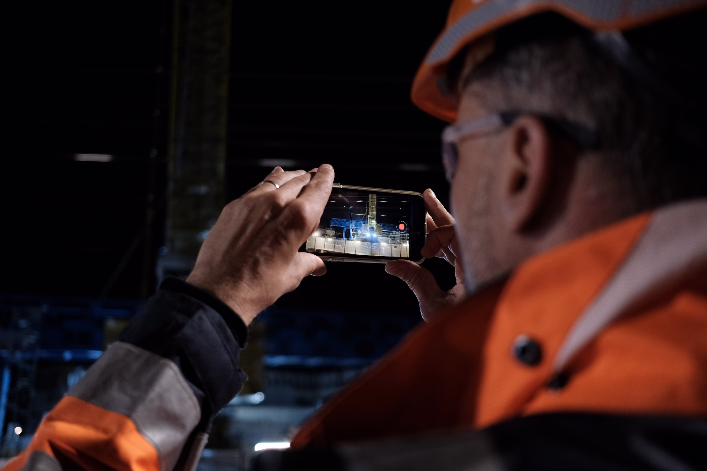 Travailleur portant un casque et gilet orange filmant un chantier industriel avec un smartphone.