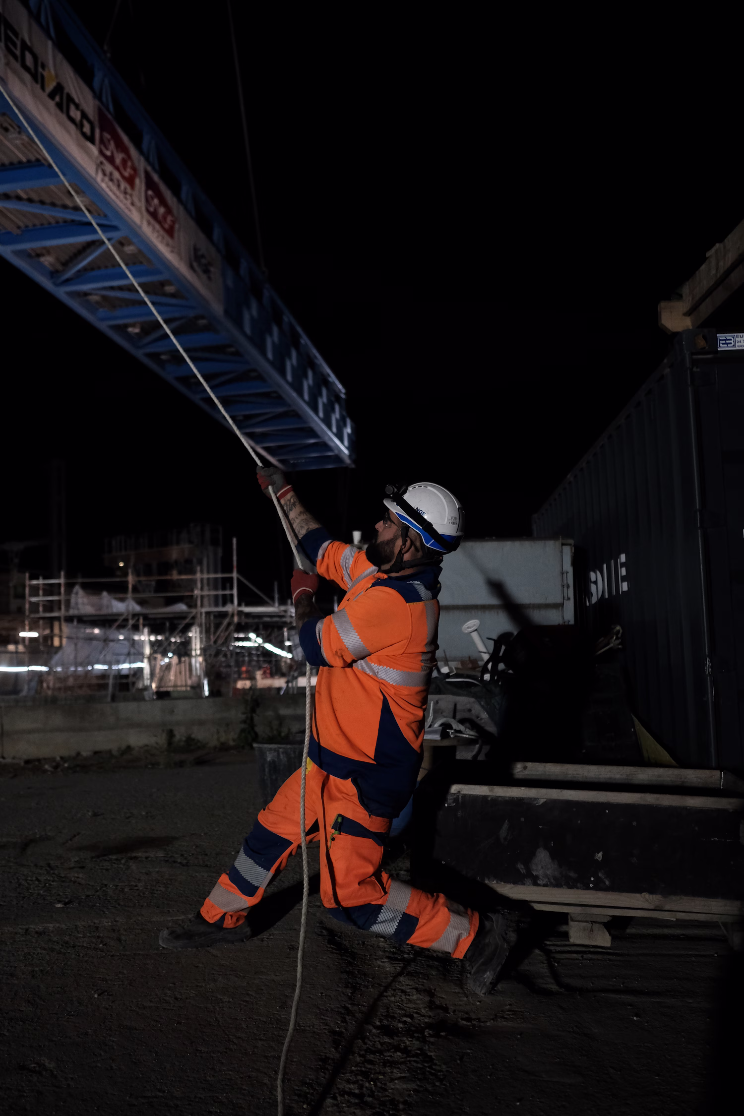 Ouvrier en tenue orange réfléchissante tirant sur une corde la nuit sur un chantier.