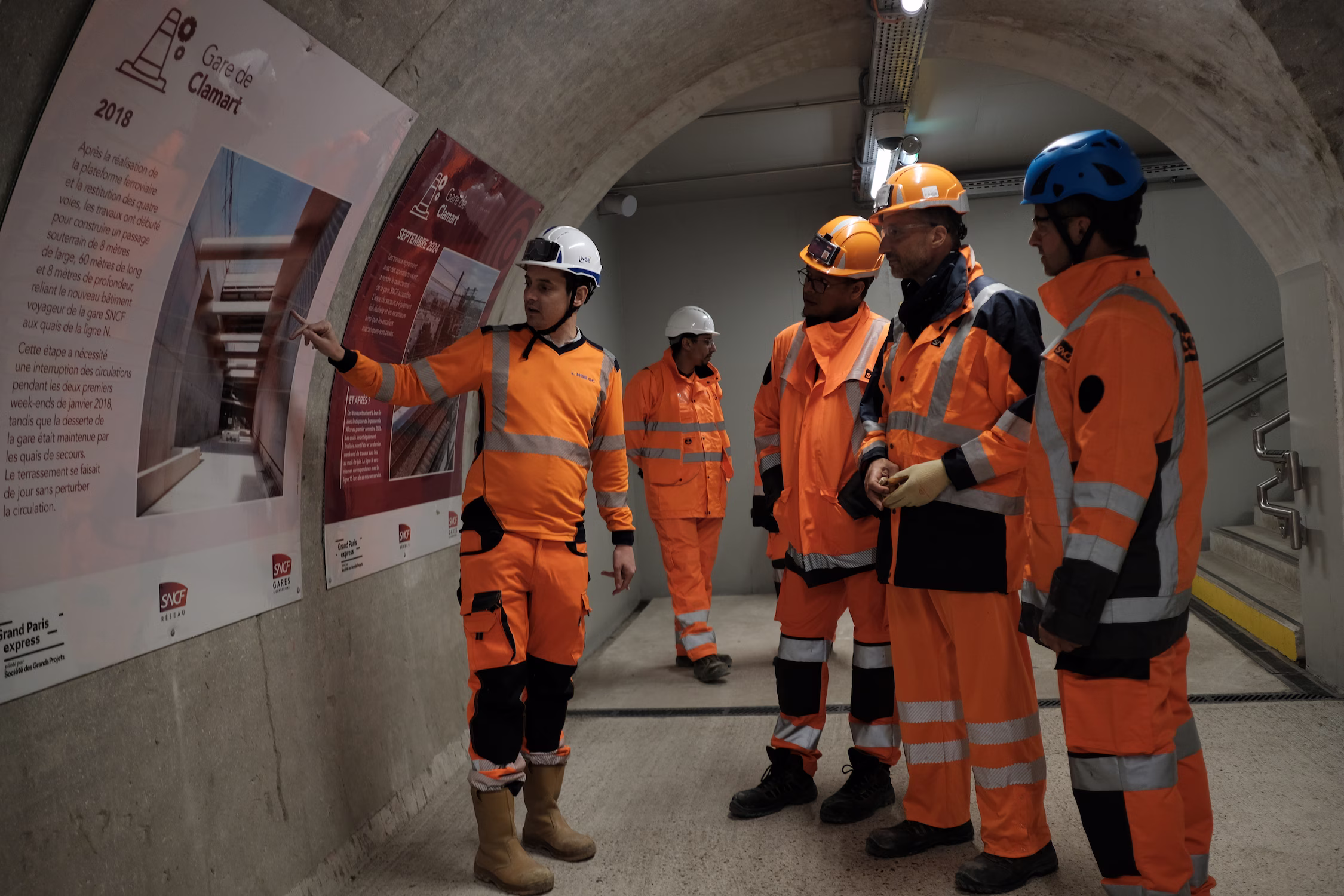 Cinq travailleurs en tenue orange regardant des panneaux explicatifs dans un tunnel en béton.