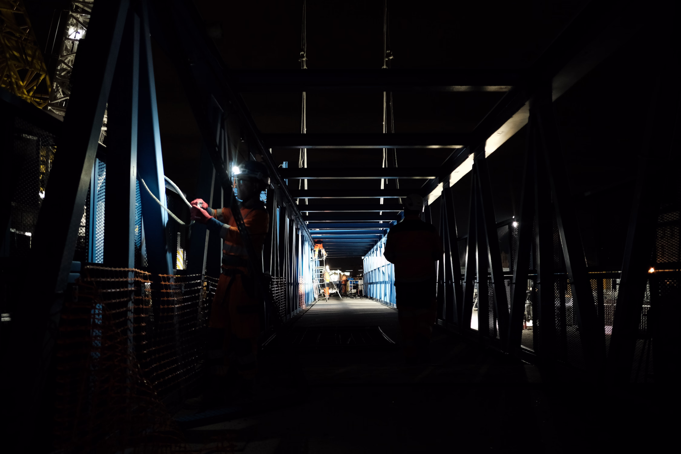 Des ouvriers portant des casques travaillent la nuit sur une passerelle métallique bleue éclairée.
