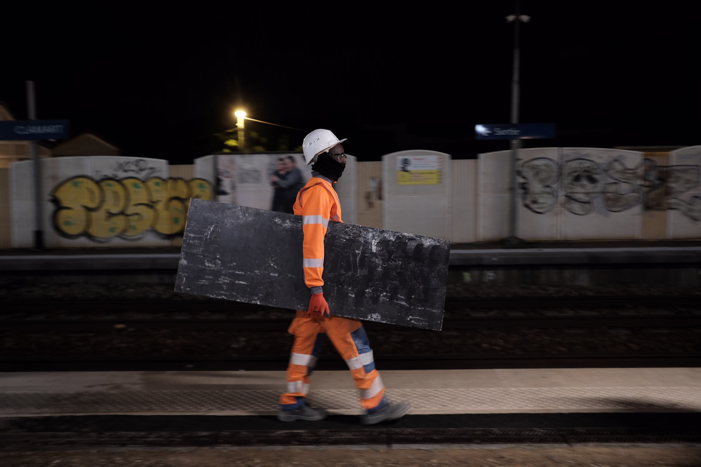 Ouvrier en tenue orange et casque portant une planche noire sur un quai de gare de nuit.