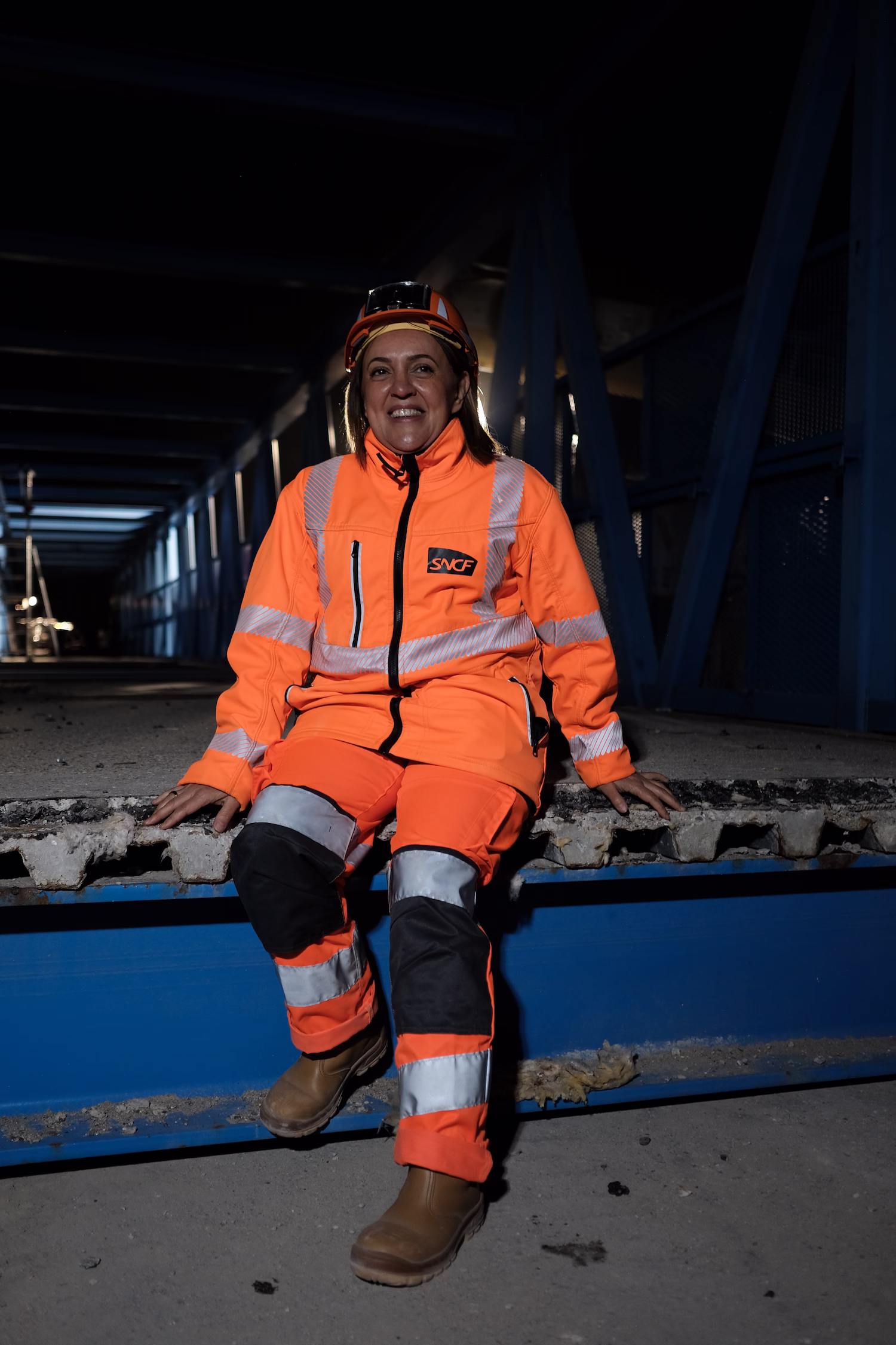 Femme en tenue haute visibilité orange SNCF souriant, assise sur une structure métallique bleue.