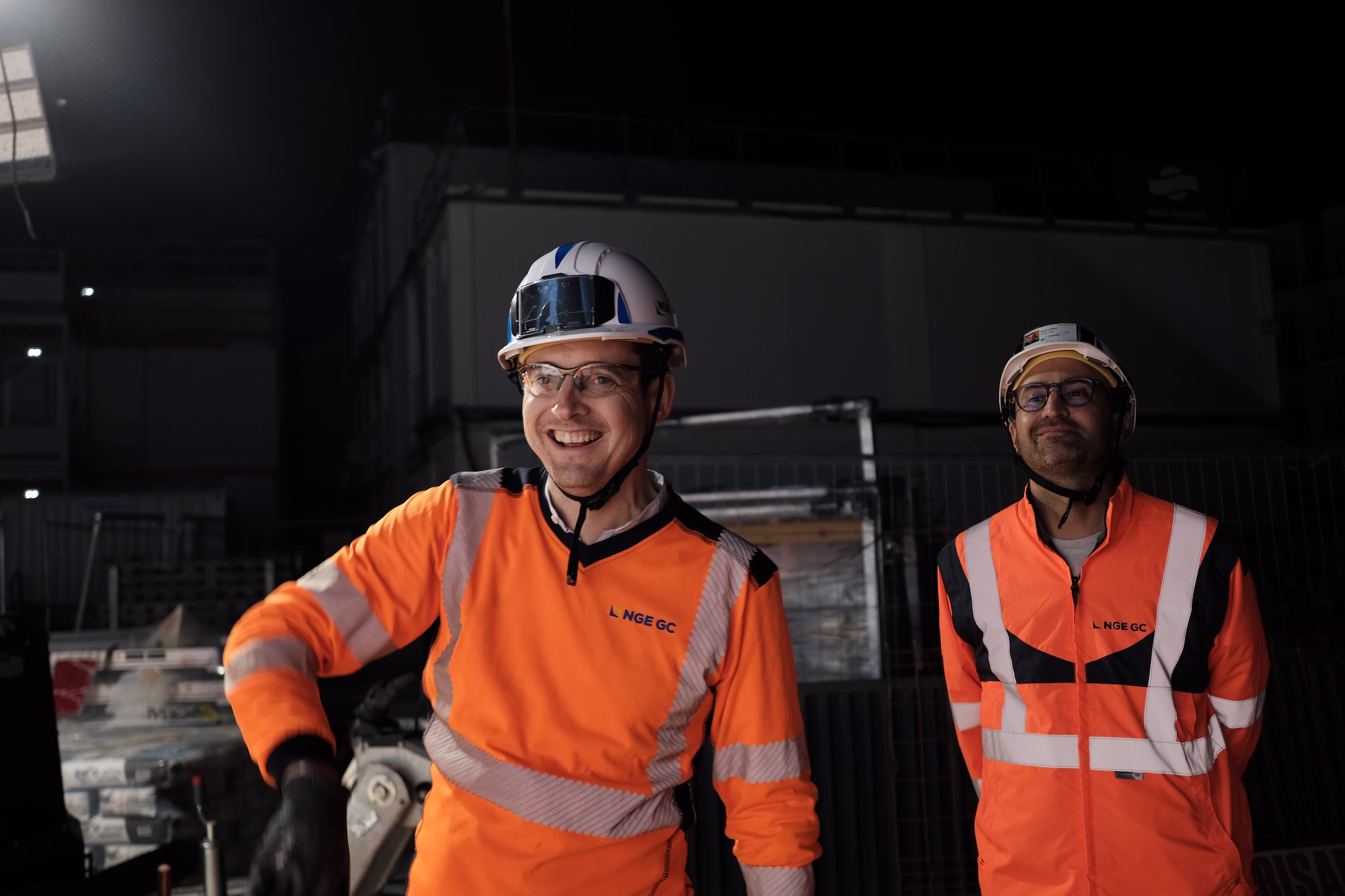 Deux ouvriers de construction portant des casques et des vestes orange NGE GC dans un chantier de nuit.
