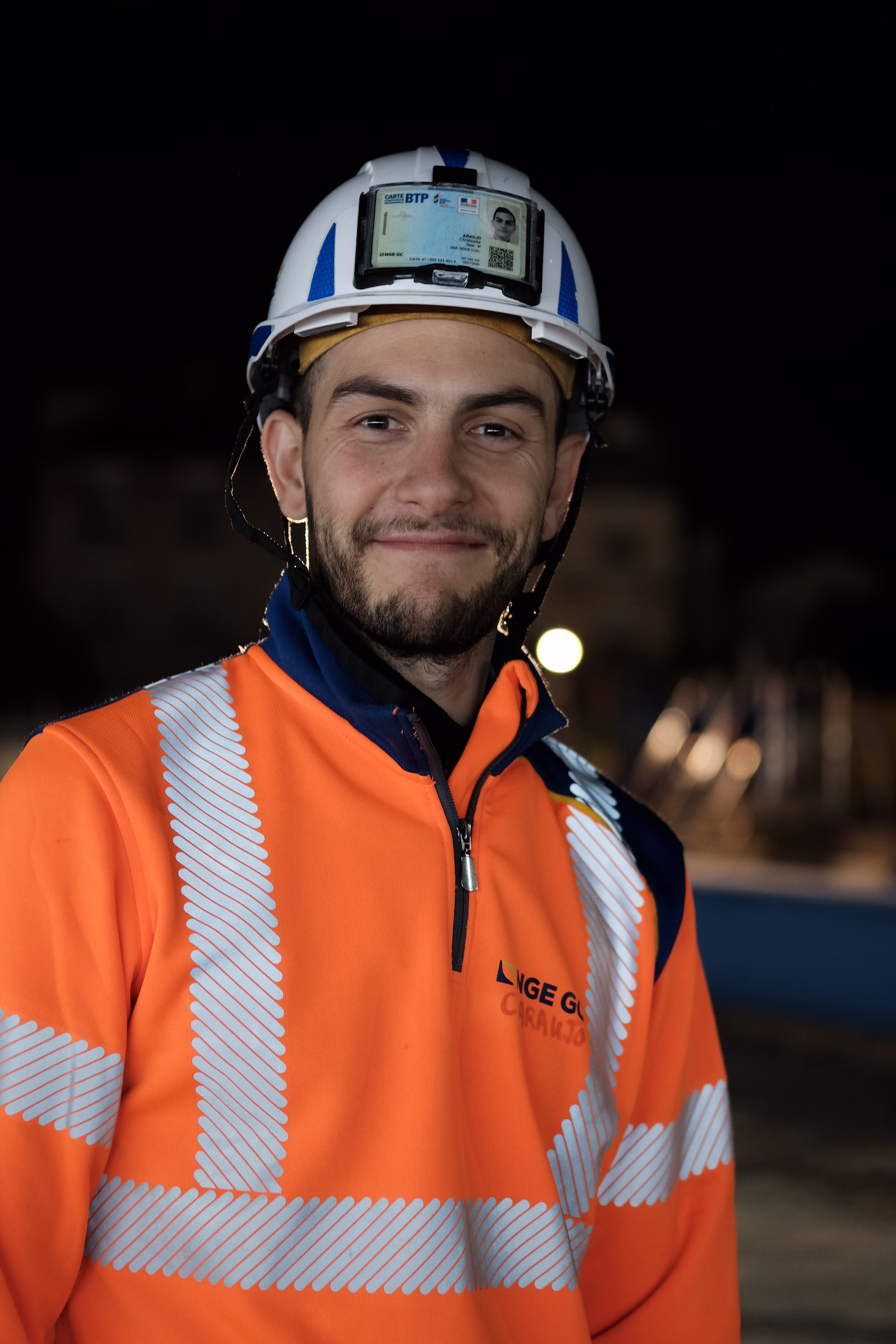 Jeune ouvrier souriant portant un casque blanc avec badge et une veste orange à bandes réfléchissantes.
