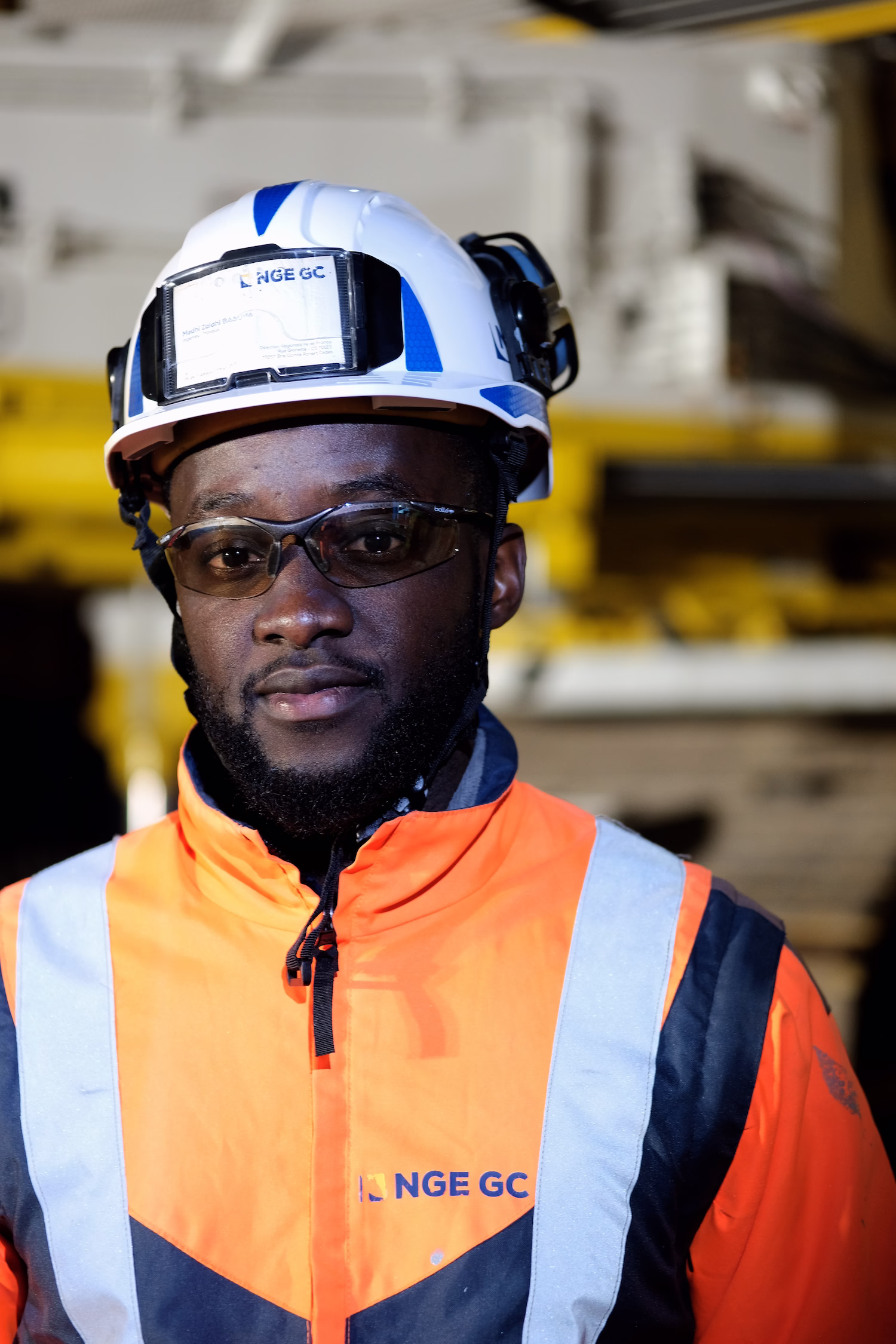 Portrait d'un travailleur portant un casque blanc et un gilet orange fluorescent avec logo NGE GC.