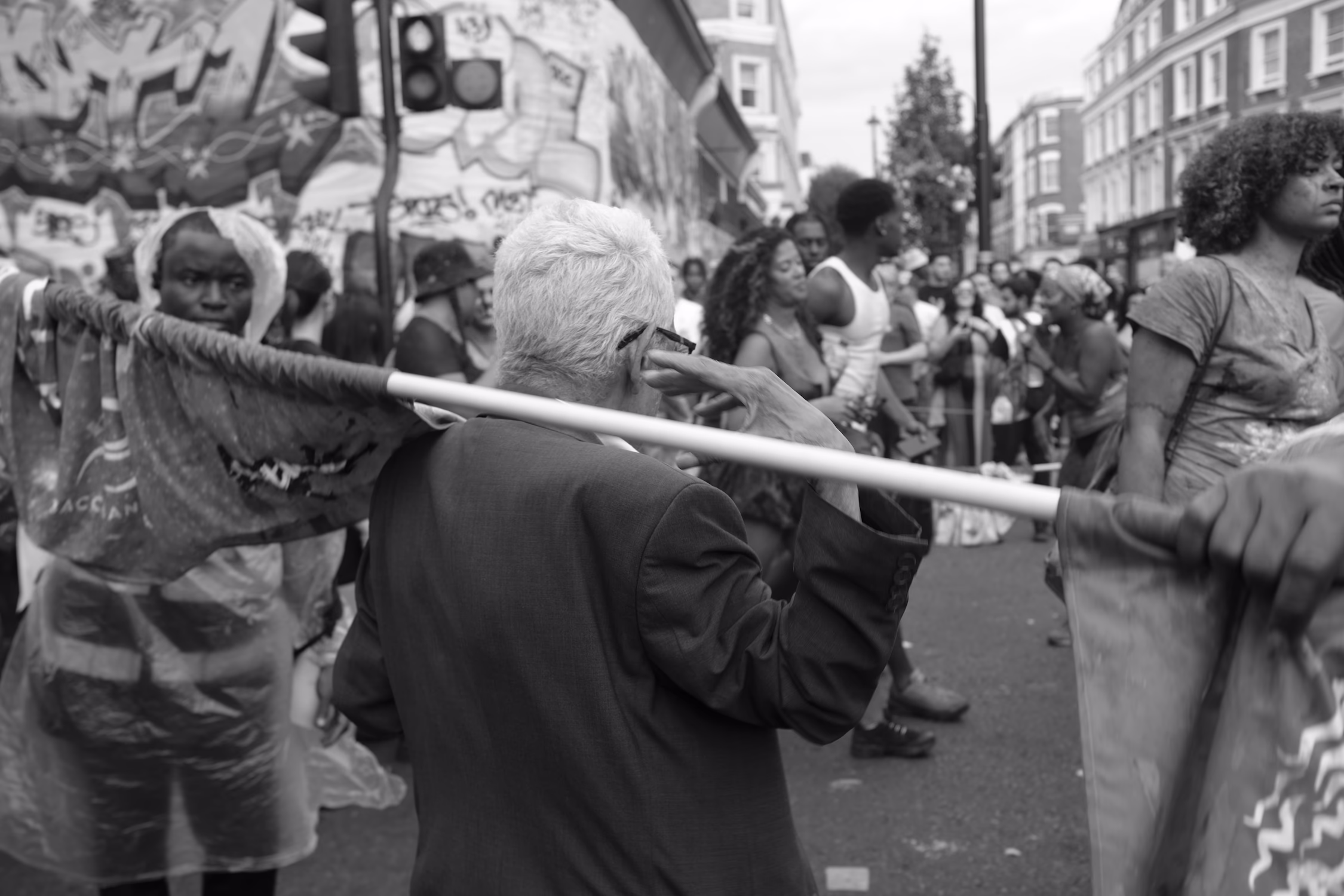 Homme vu de dos tenant une perche lors d'un carnaval animé en pleine rue avec foule en arrière-plan.