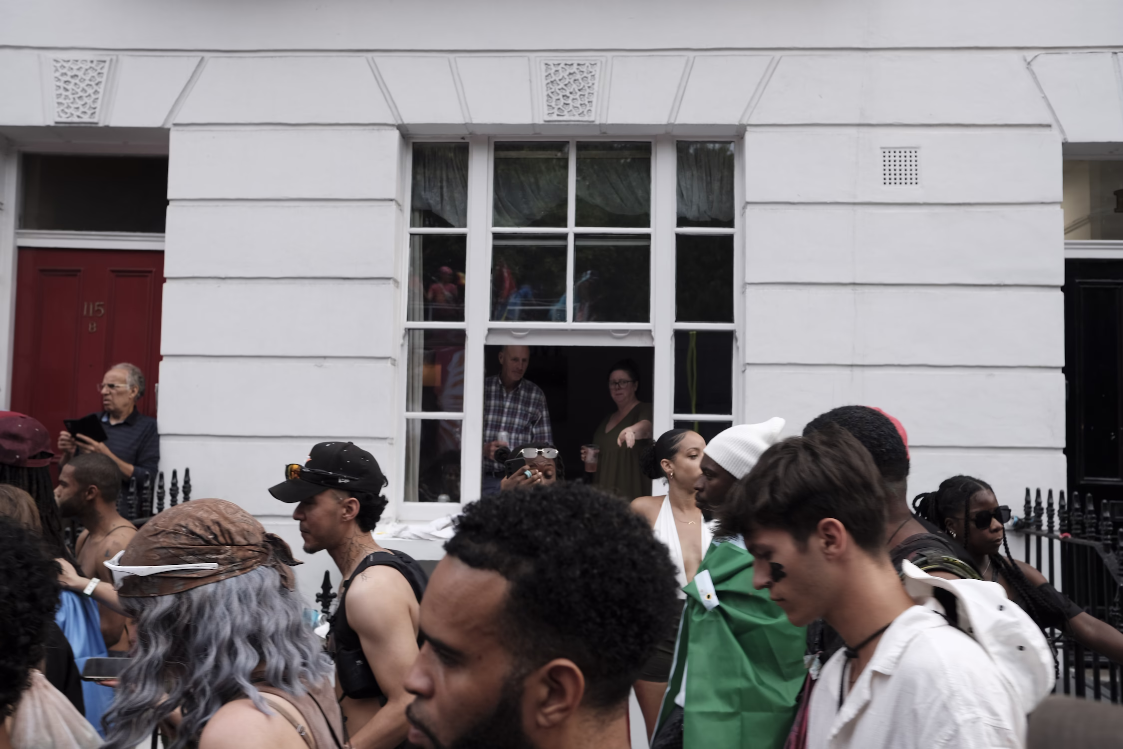 Défilé du carnaval devant le mur d'une façade blanche avec deux personnes regardant par une fenêtre ouverte.