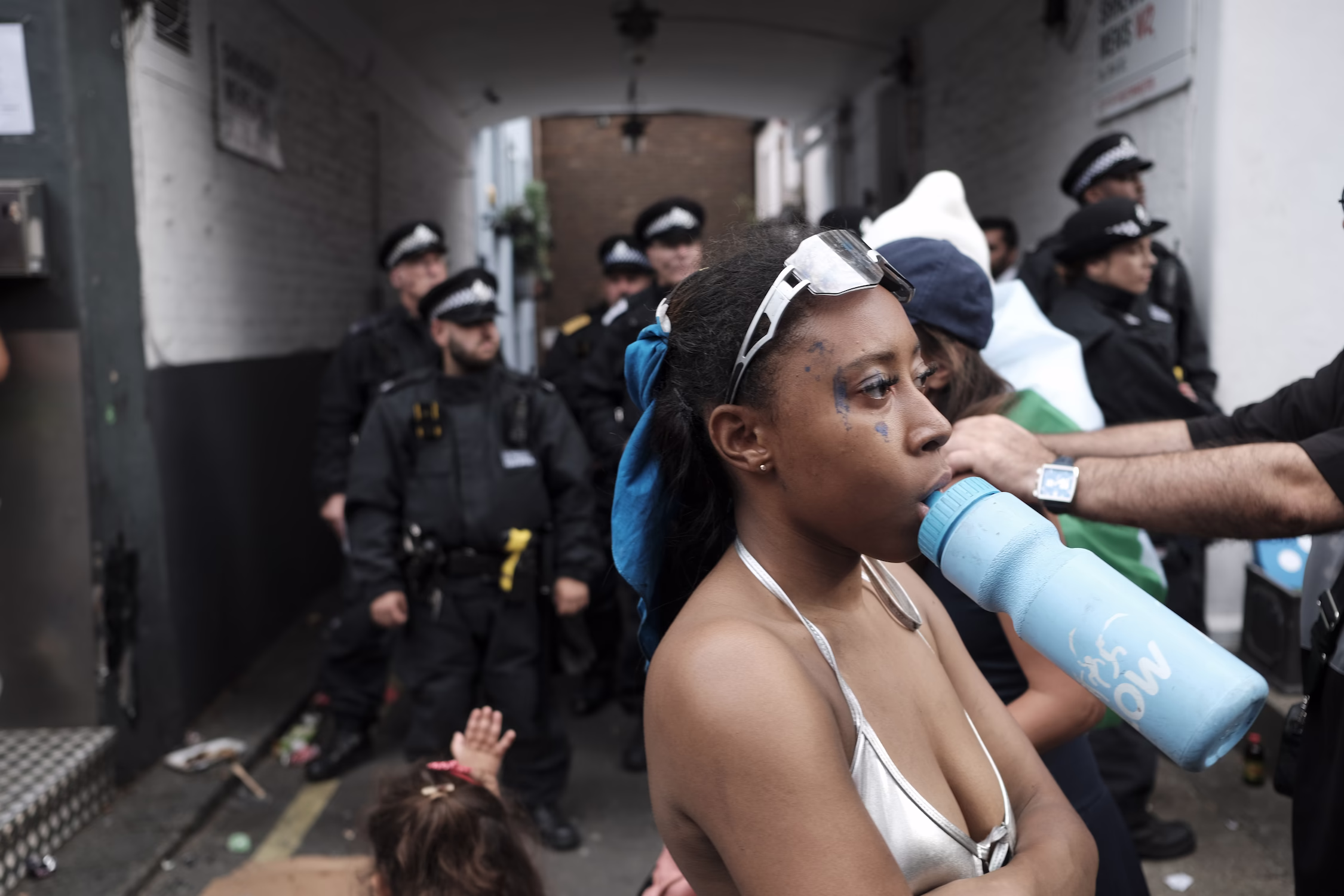 Femme buvant dans une gourde bleue devant des policiers en uniforme dans une ruelle.