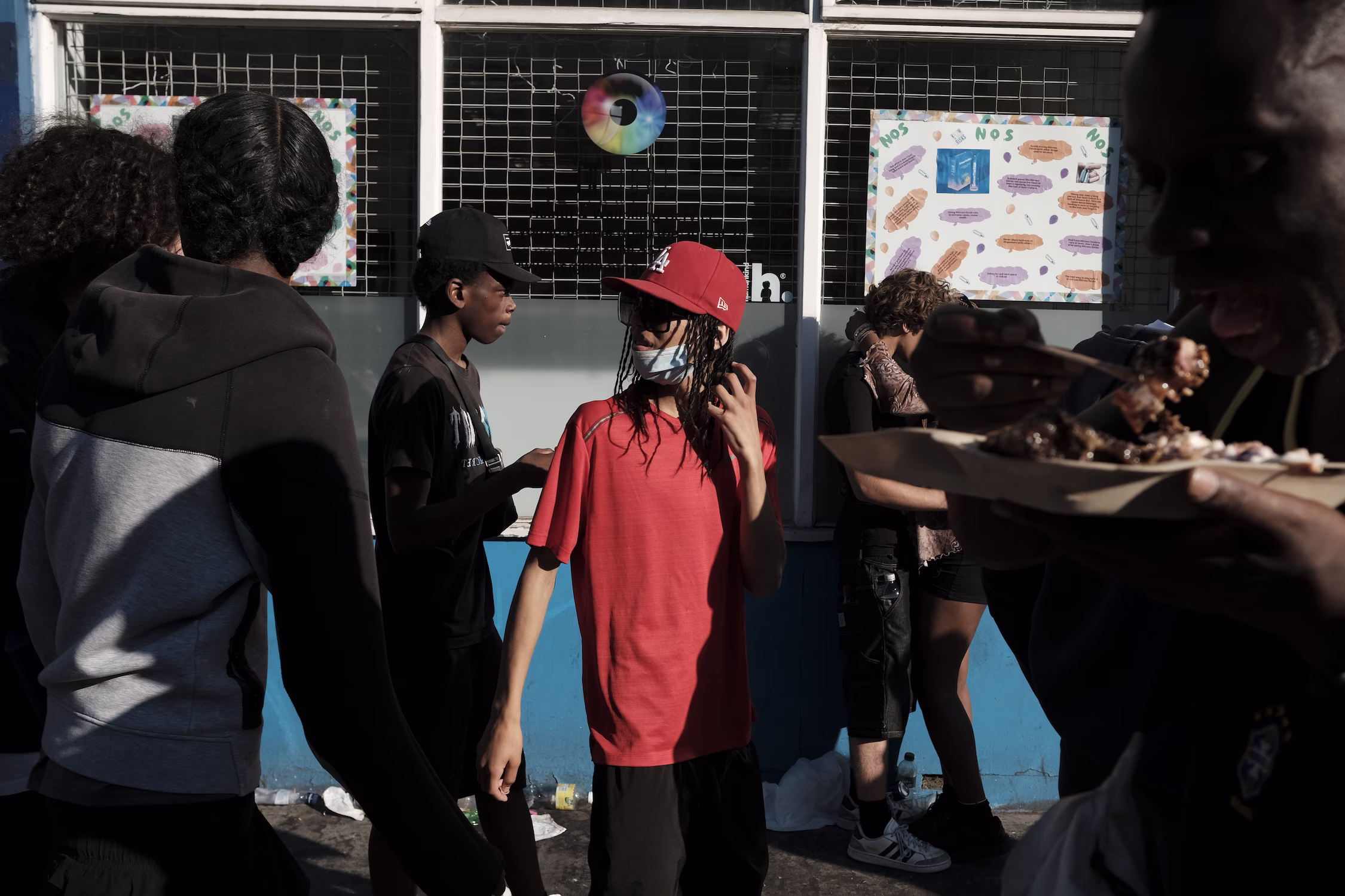Groupe de jeunes debout devant un mur avec des affiches colorées, une personne mange au premier plan.